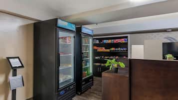 A hotel lobby with two large black refrigerators, a digital kiosk, and a reception desk with a potted plant.