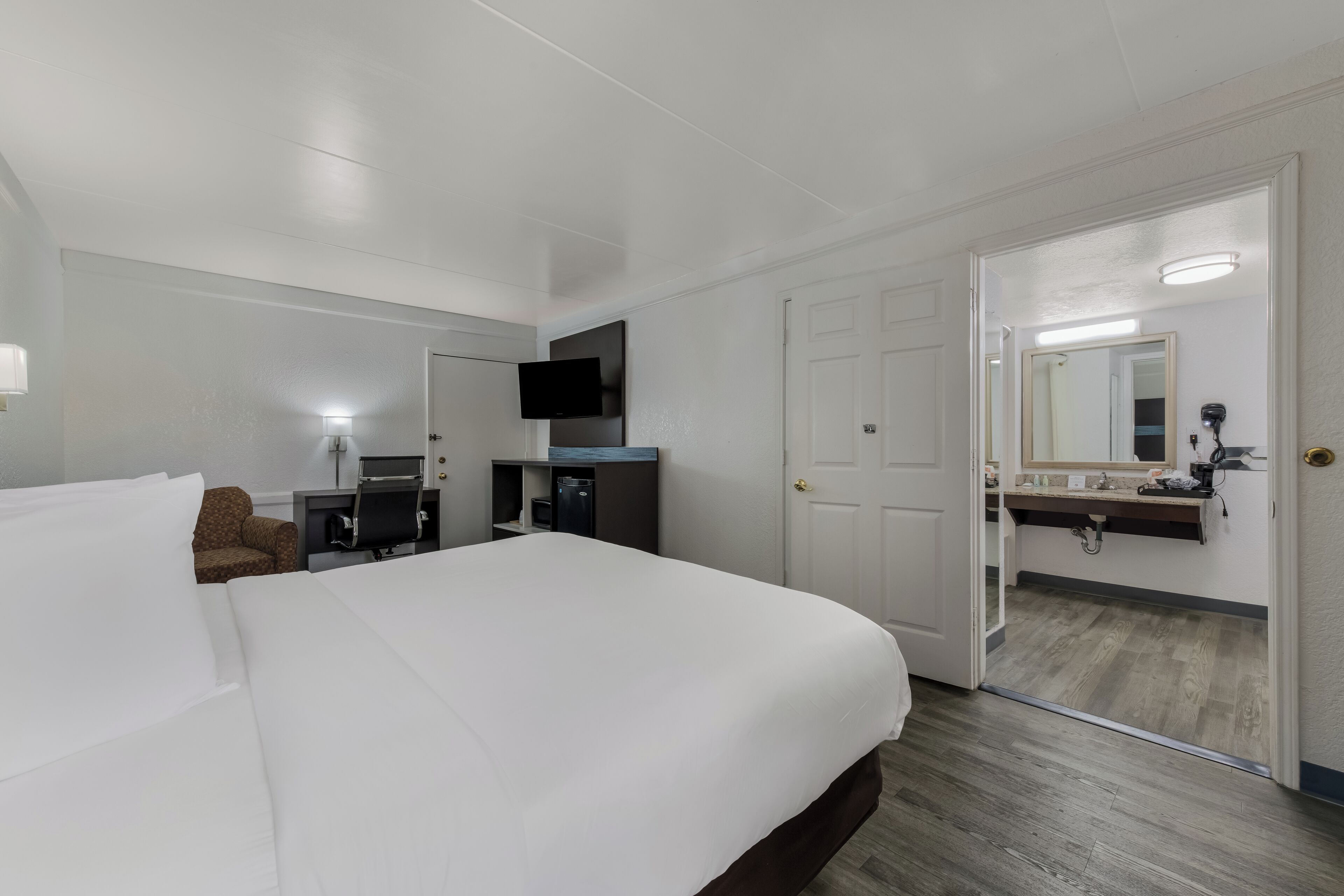 A hotel room with a large bed, a desk with a chair, a flat-screen TV mounted on the wall, and a bathroom with a mirror and sink.