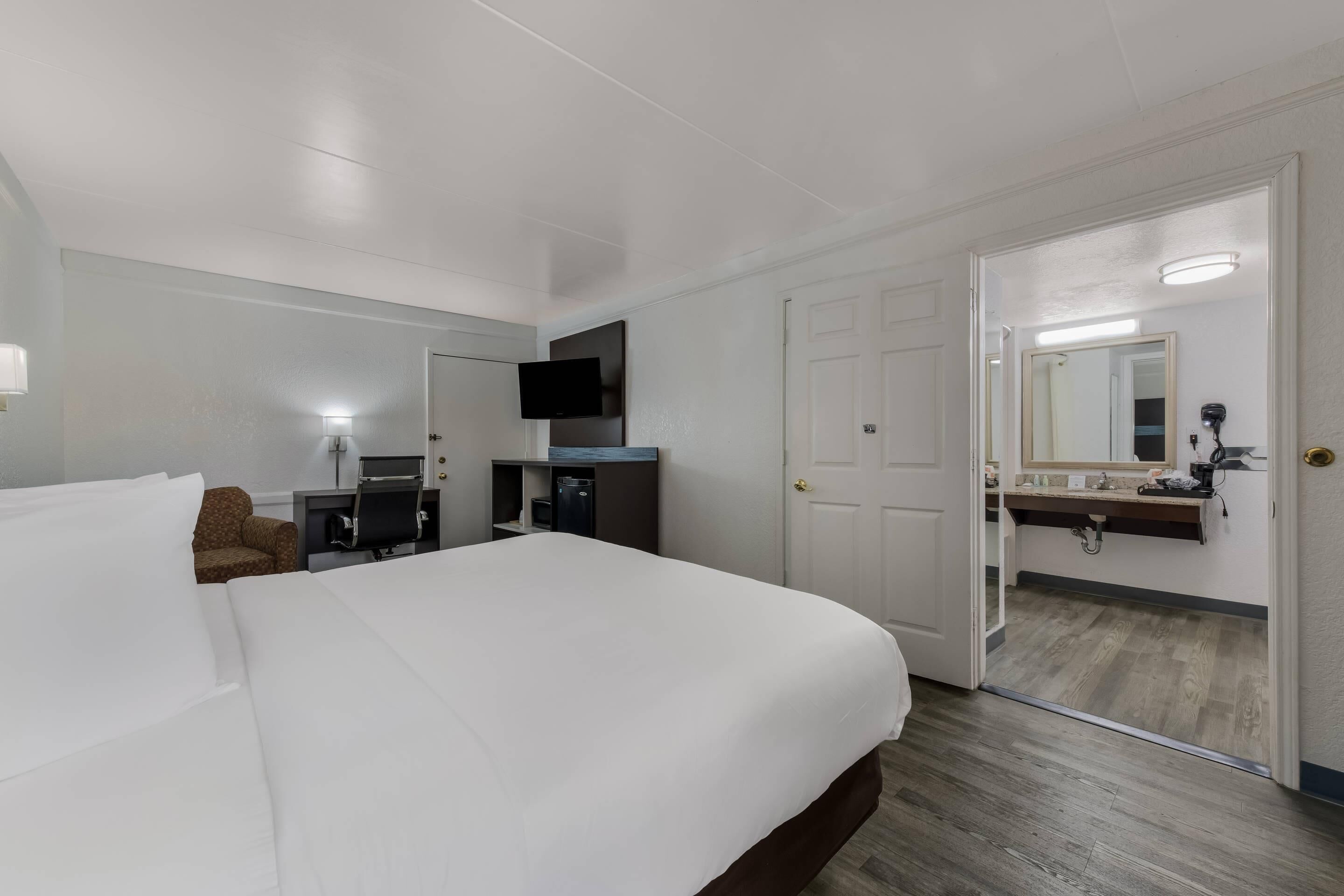 A hotel room with a large bed, a desk with a chair, a flat-screen TV, a mirror, and a door leading to a bathroom.