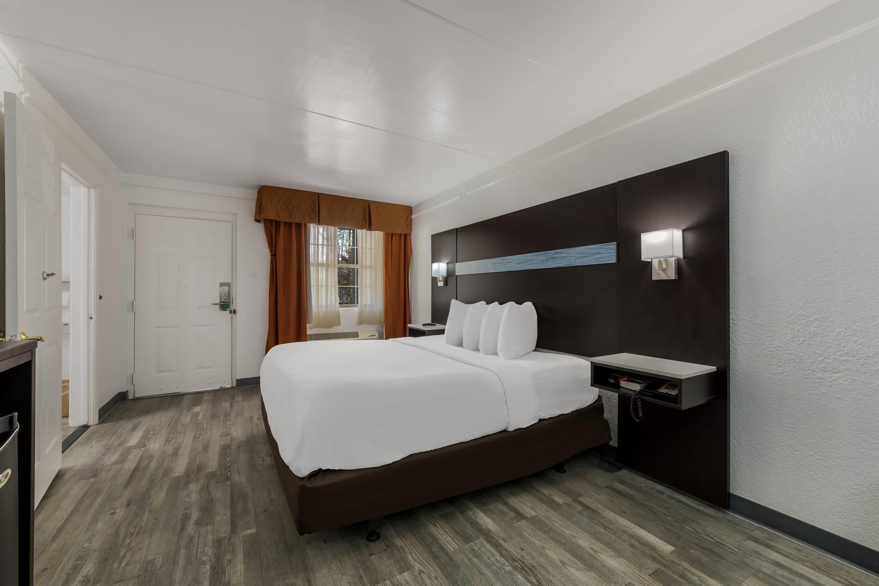 A hotel room with a large bed, a bedside table, a nightstand, a wall-mounted lamp, and a window with curtains.