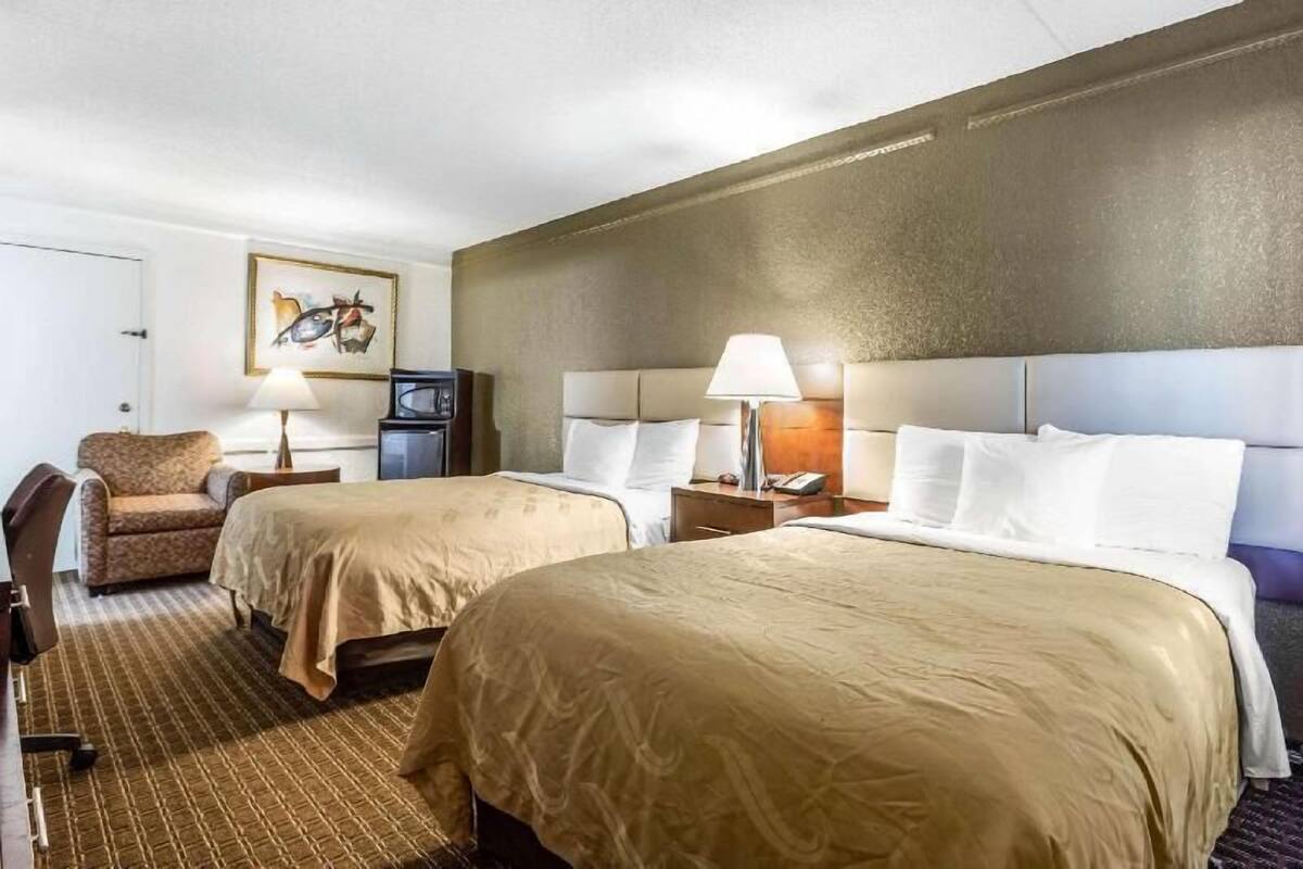 Ein Hotelzimmer mit zwei Betten, einem Sessel, einem Schreibtisch, einer Lampe und einem Fernseher.