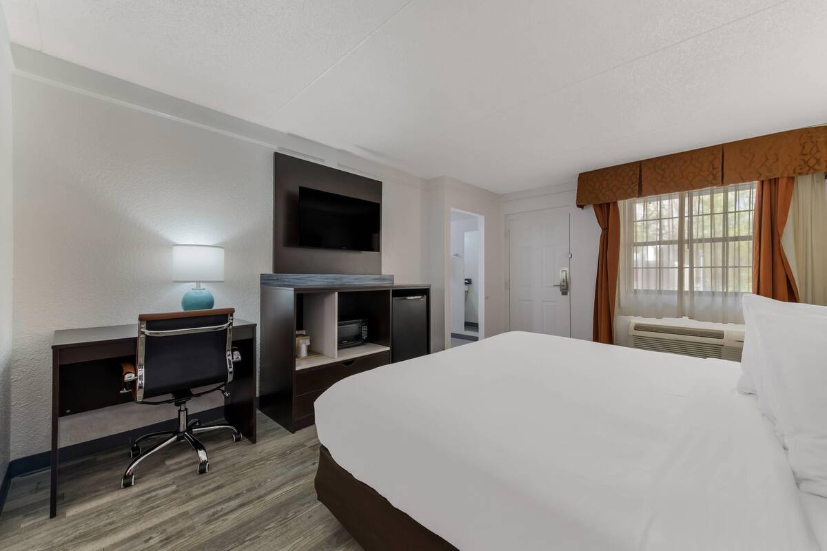 Ein Hotelzimmer mit einem großen Bett, einem Schreibtisch mit Stuhl, einem Flachbildfernseher, einer Mikrowelle und einem Fenster mit Vorhängen.