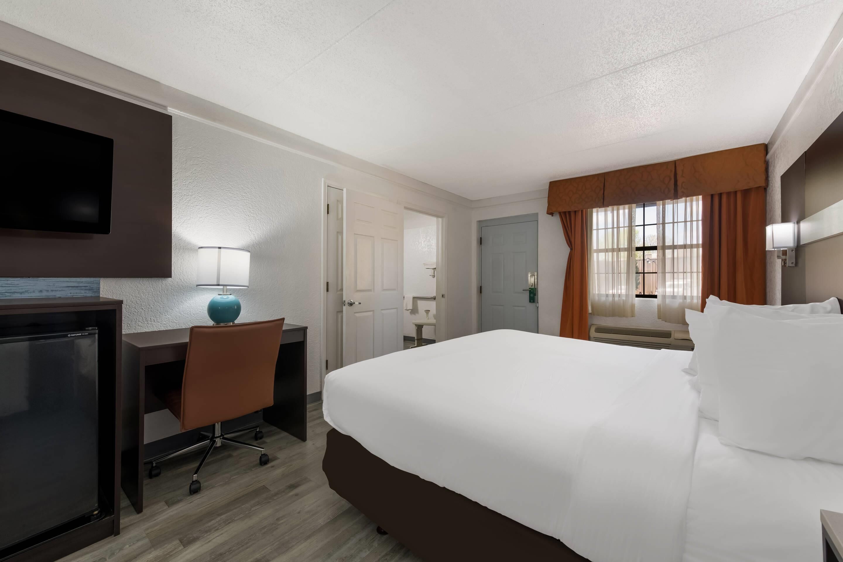 A hotel room with a bed, desk, chair, TV, lamp, and a window with curtains.