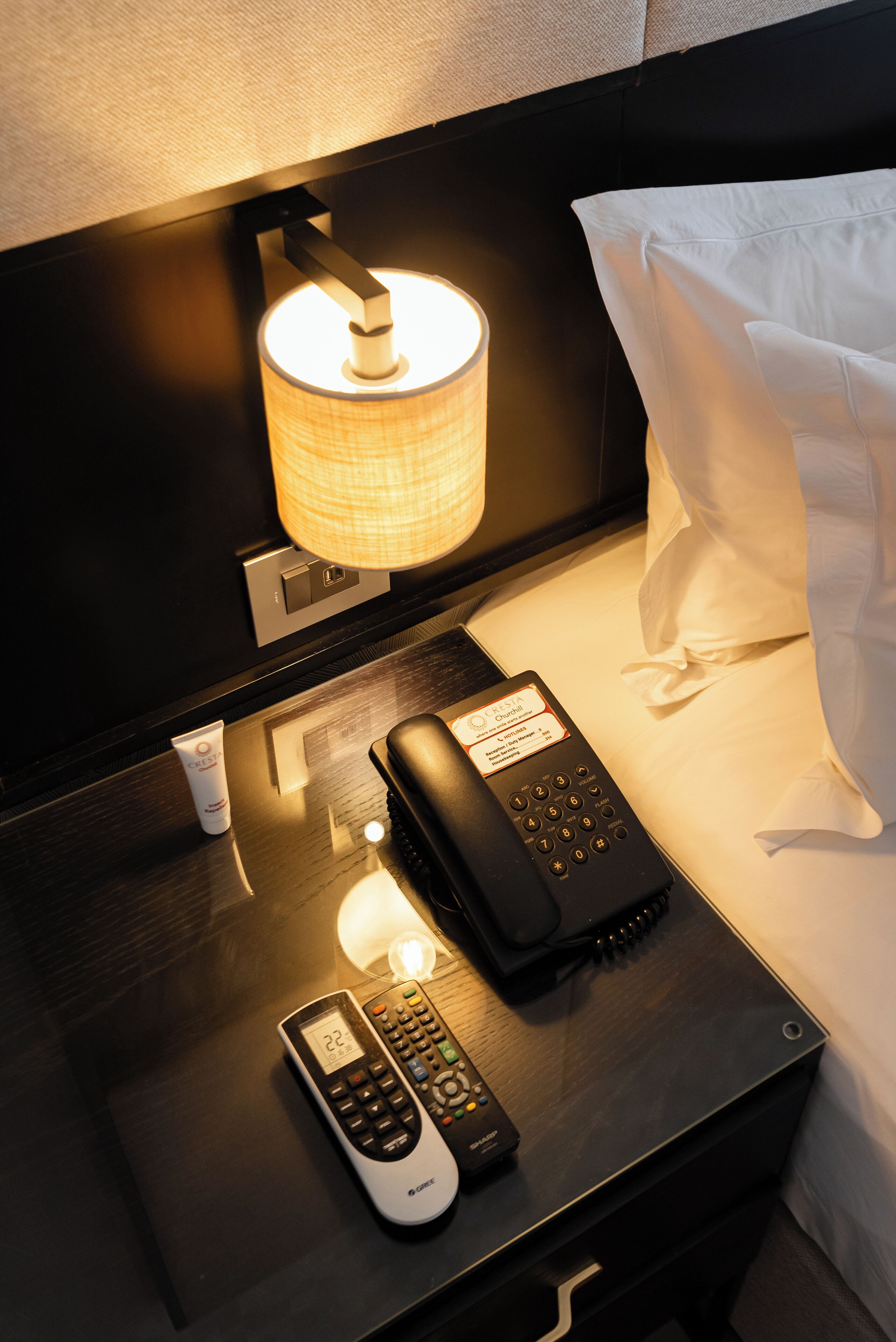 A hotel room with a bedside table featuring a lamp, a remote control, a phone, and a tube of cream.
