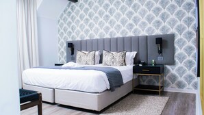 A modern bedroom with a grey upholstered bed, white bedding, patterned pillows, a bedside table with a lamp, and a patterned wallpaper. - Cresta Churchill (Bulawayo)