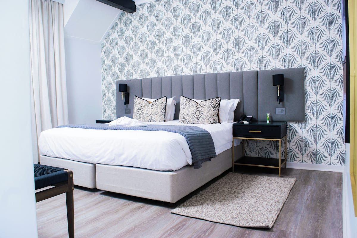 A modern bedroom with a grey upholstered bed, white bedding, patterned pillows, a bedside table with a lamp, and a patterned wallpaper.