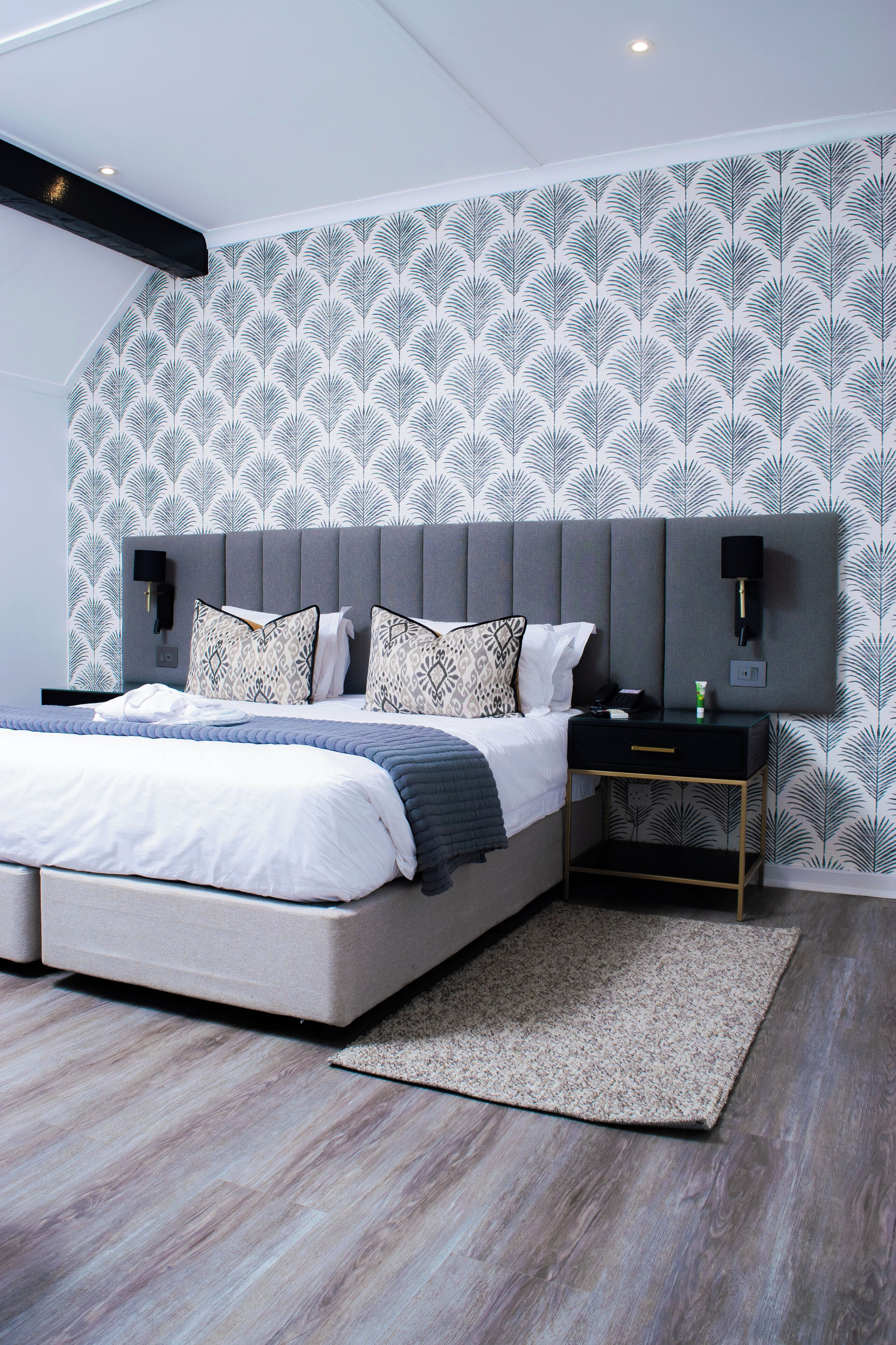 A modern bedroom with a large bed, a grey upholstered headboard, a bedside table with a lamp, and patterned wallpaper.