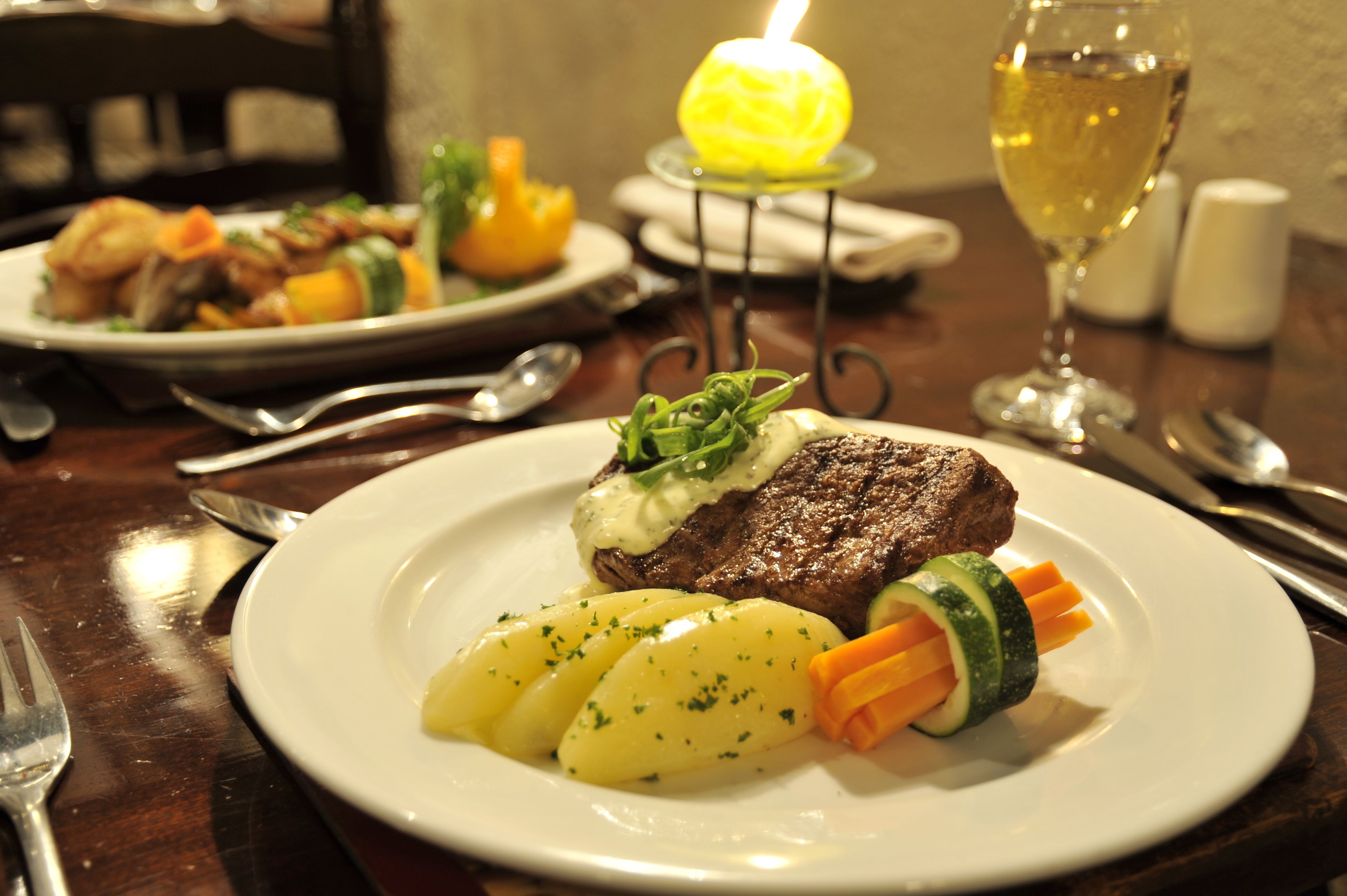 A plate with steak, potatoes, and vegetables, and a glass of white wine on a table.