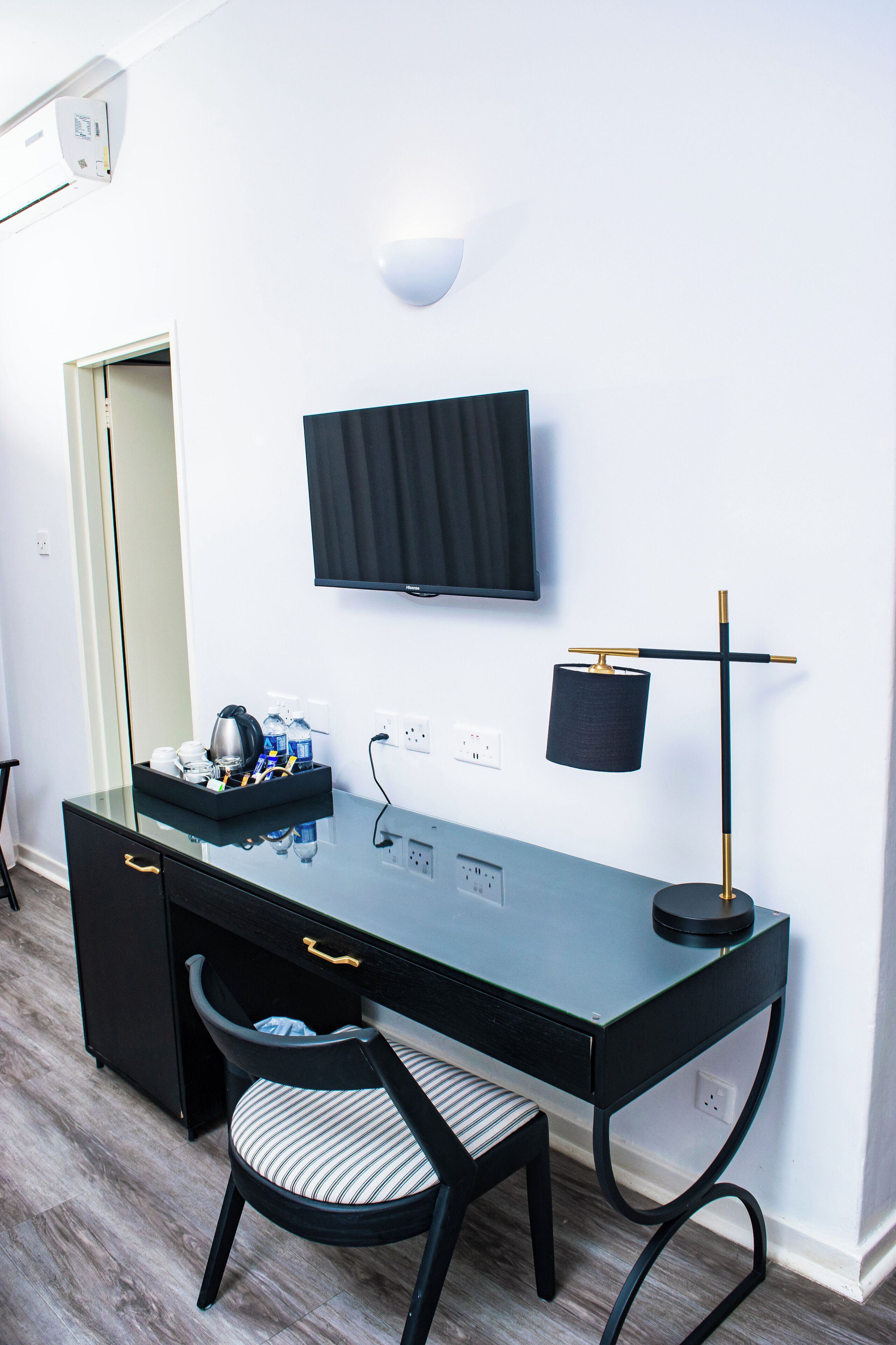 A modern room with a glass desk, a black chair with striped cushion, a mounted flat-screen TV, and a desk lamp.