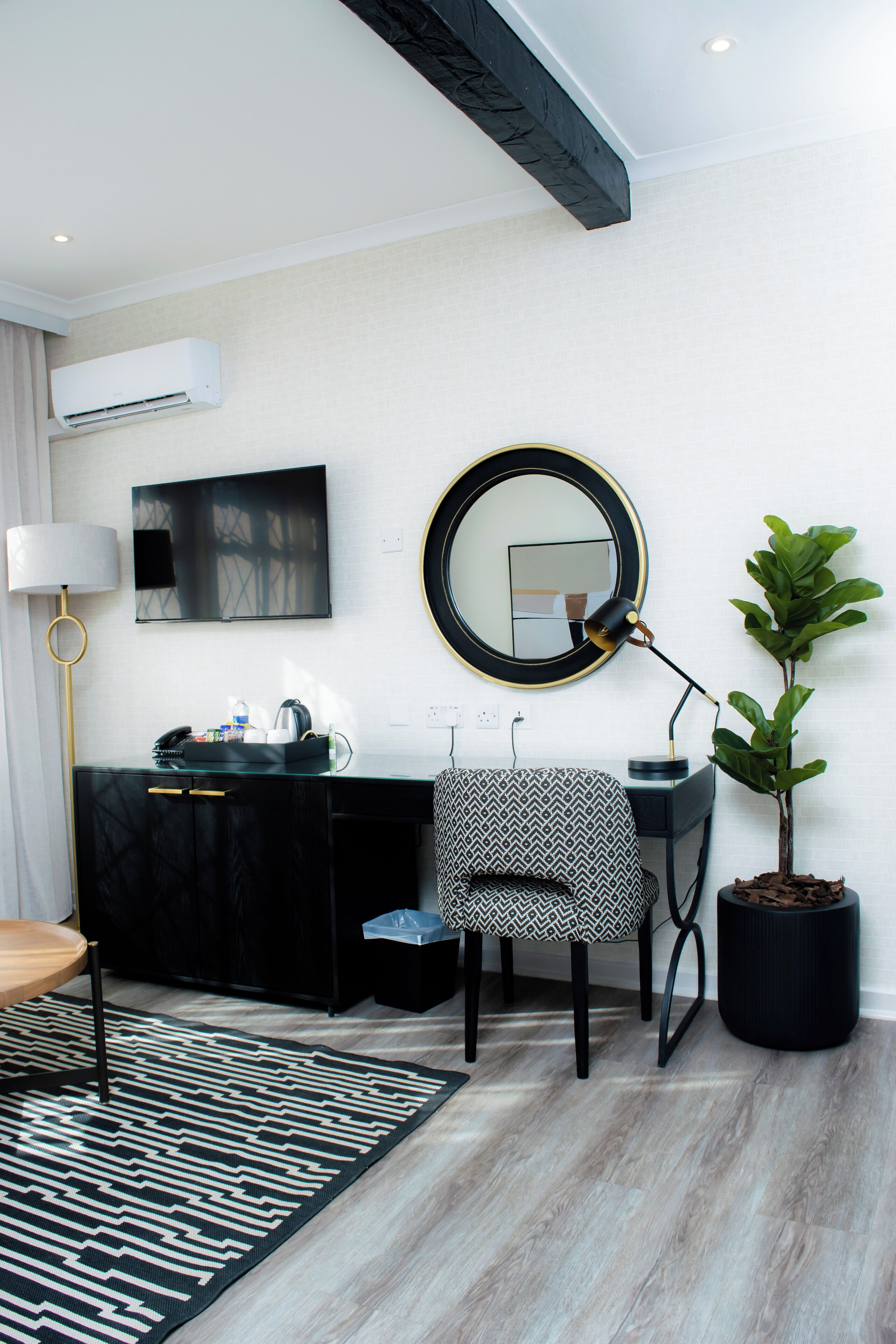 Une chambre d’hôtel moderne avec un canapé à motifs noir et blanc, un miroir rond, une plante en pot et un bureau avec une lampe.