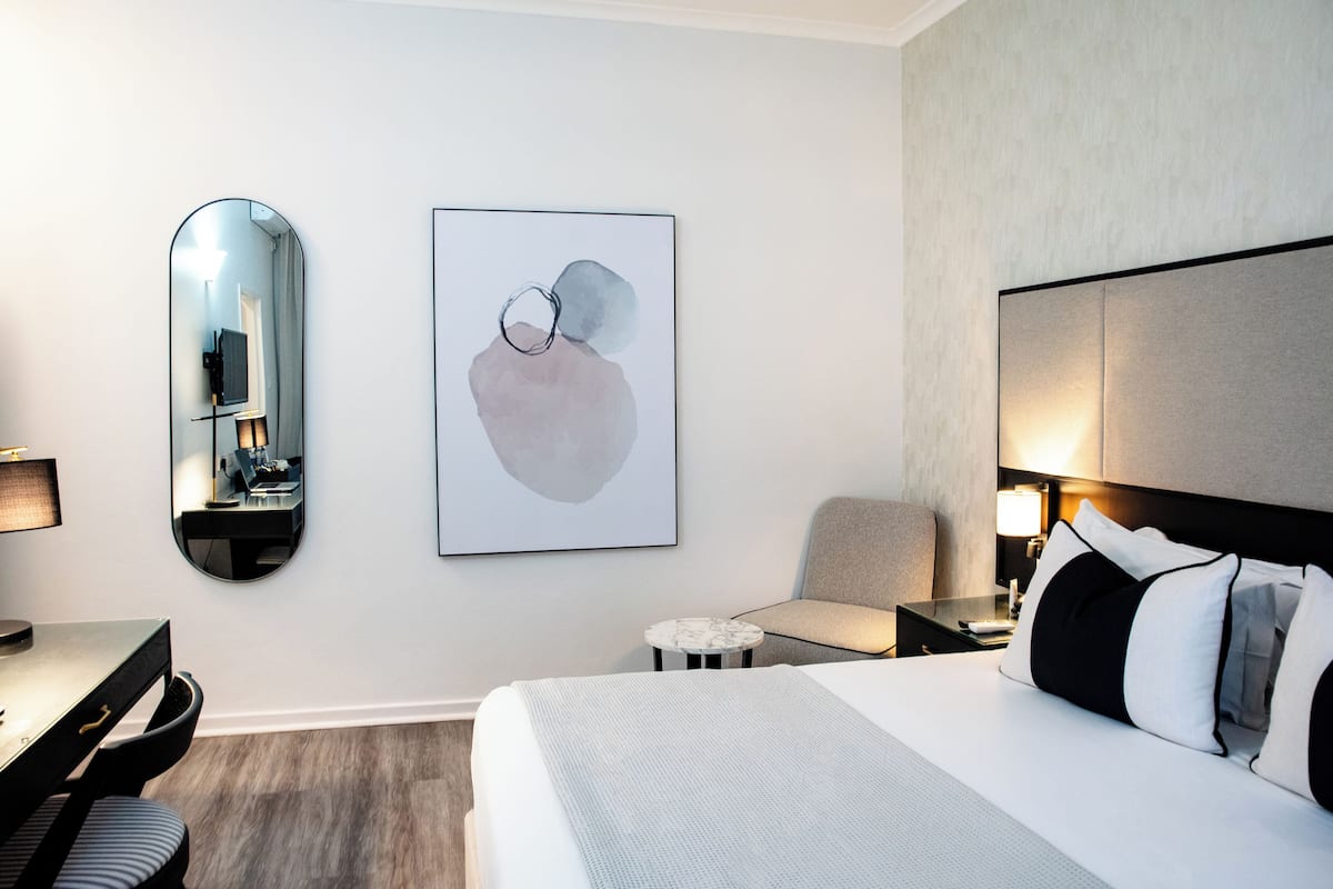 A modern hotel room with a bed, a desk, a chair, a round table, a mirror, and artwork on the wall.