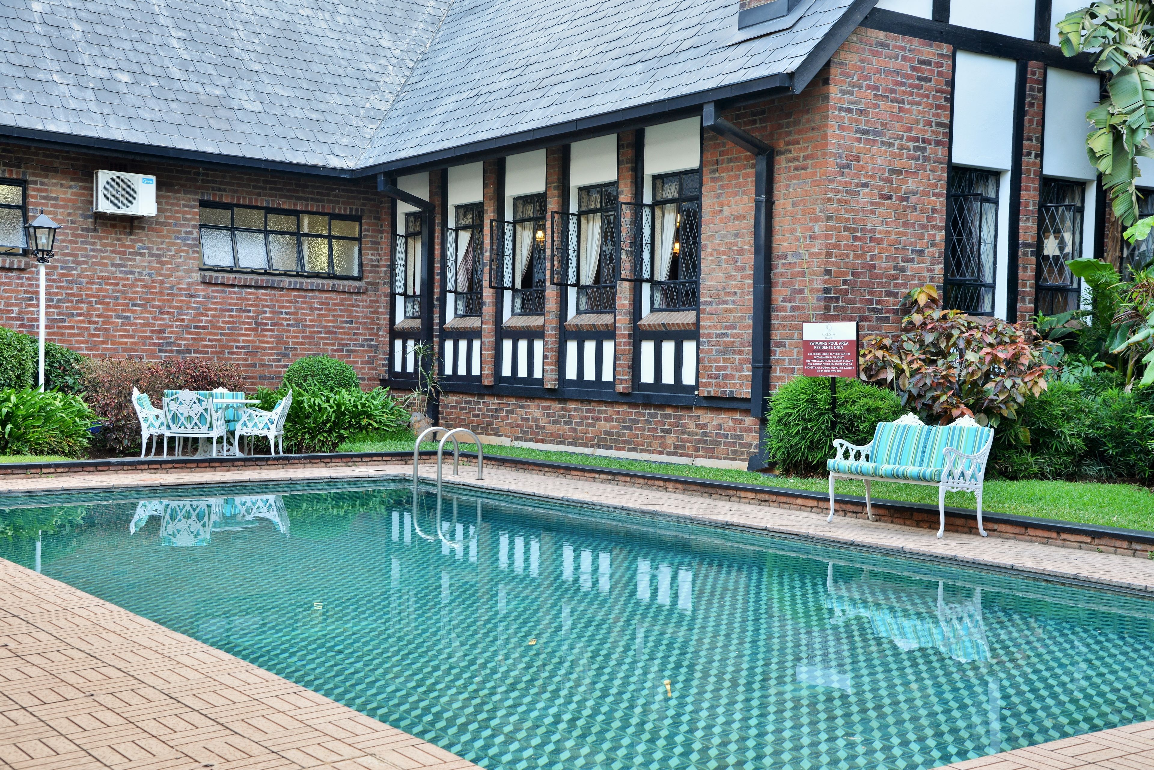 A swimming pool with a tiled deck, a brick building with large windows, and outdoor seating.