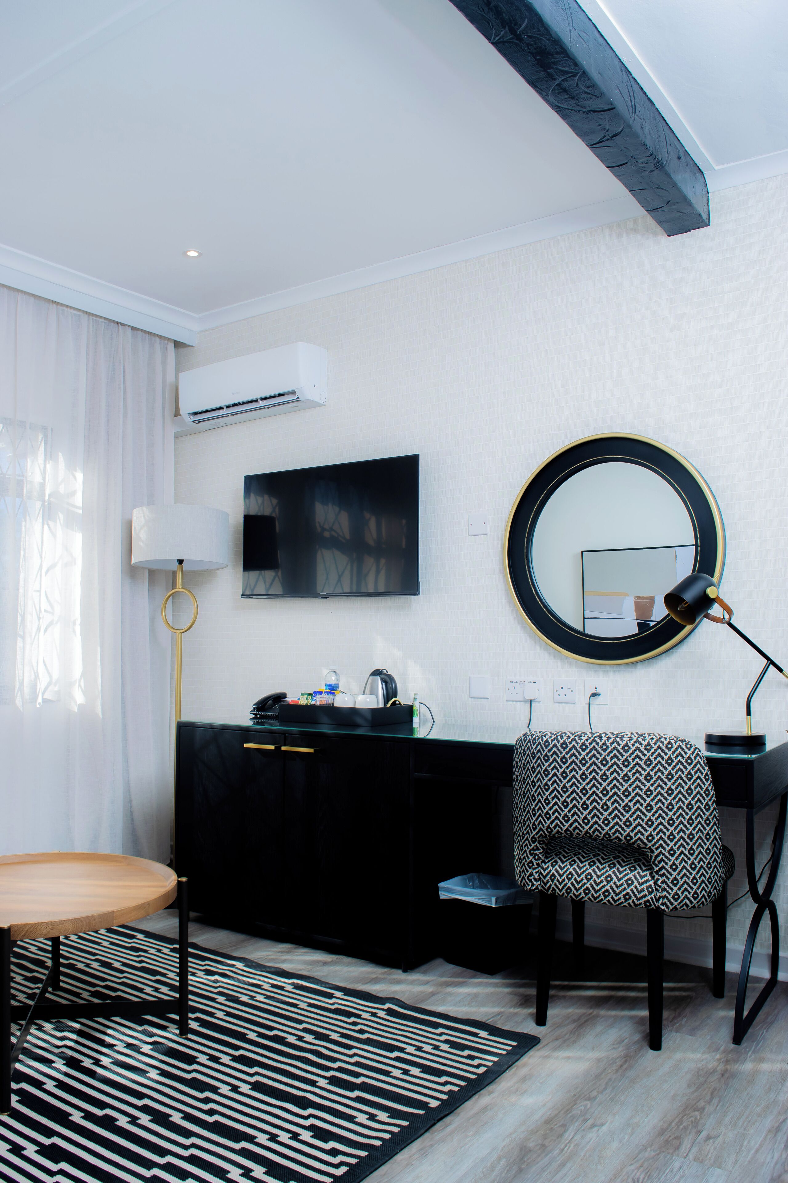 A modern hotel room with a black and white patterned chair, a round mirror with a gold frame, a black desk, a round table, and a black and white patterned rug.