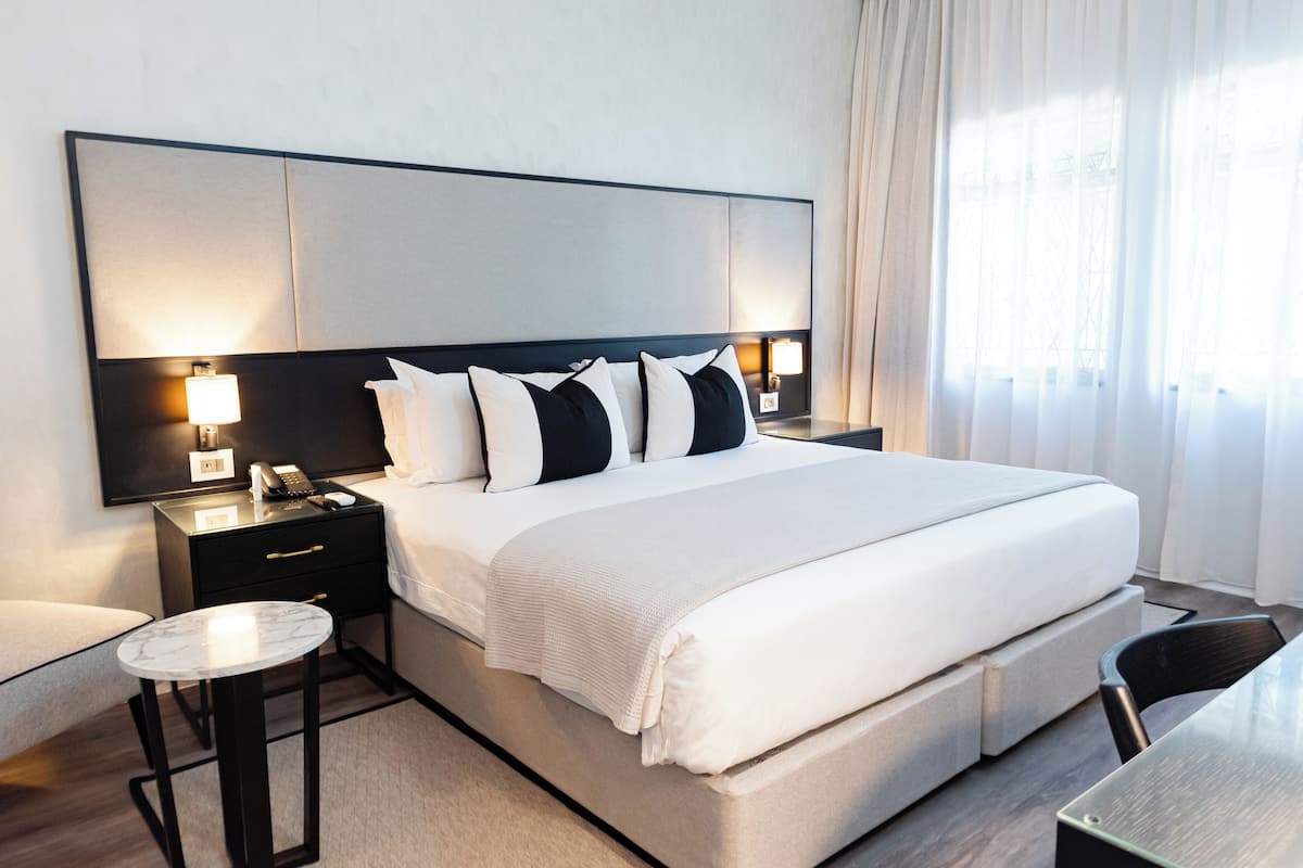 A modern hotel room with a large bed, bedside tables with lamps, a small round table, and a chair.