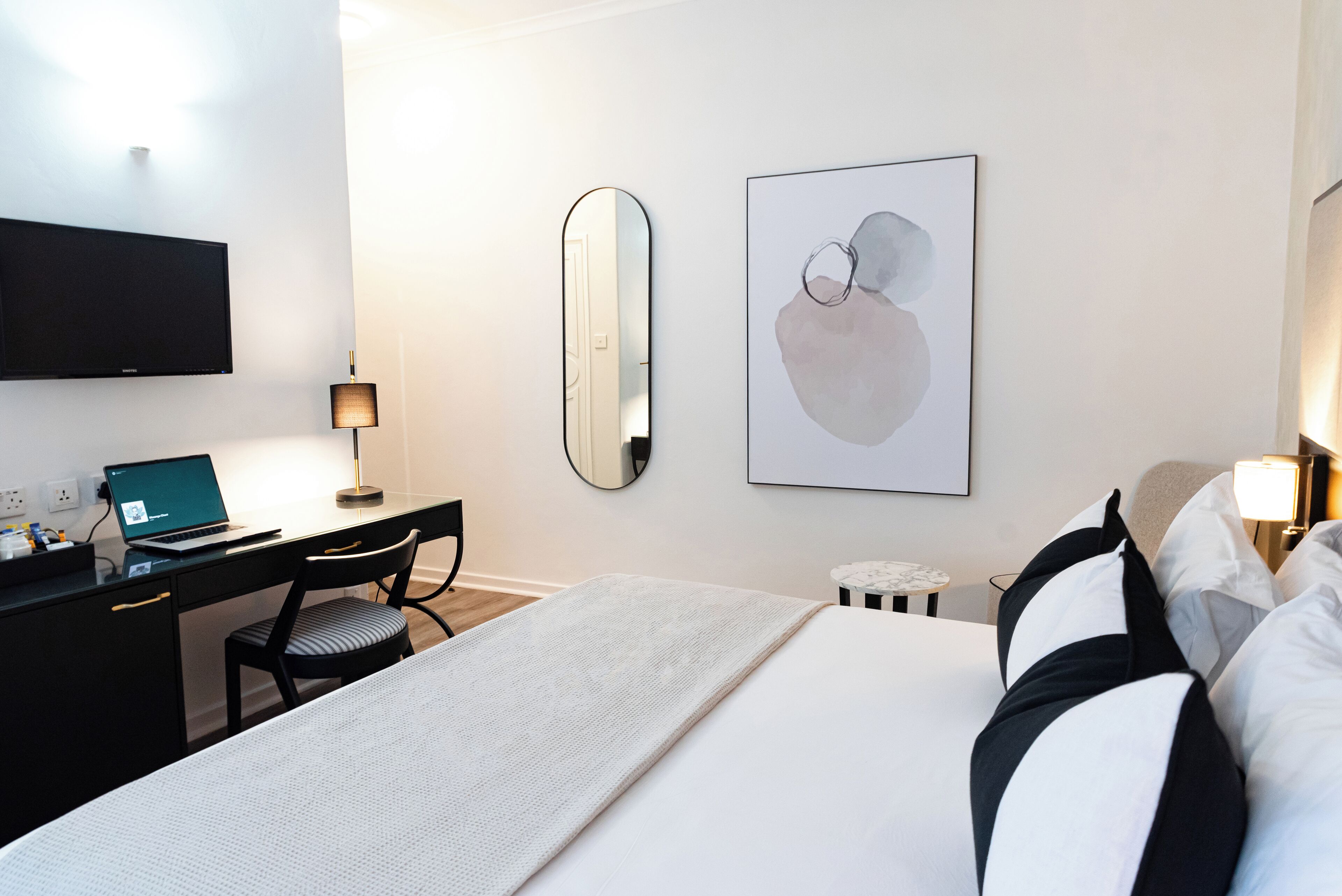 Habitación de hotel con cama, escritorio con ordenador portátil, silla, televisor montado en la pared, espejo y una obra de arte enmarcada.