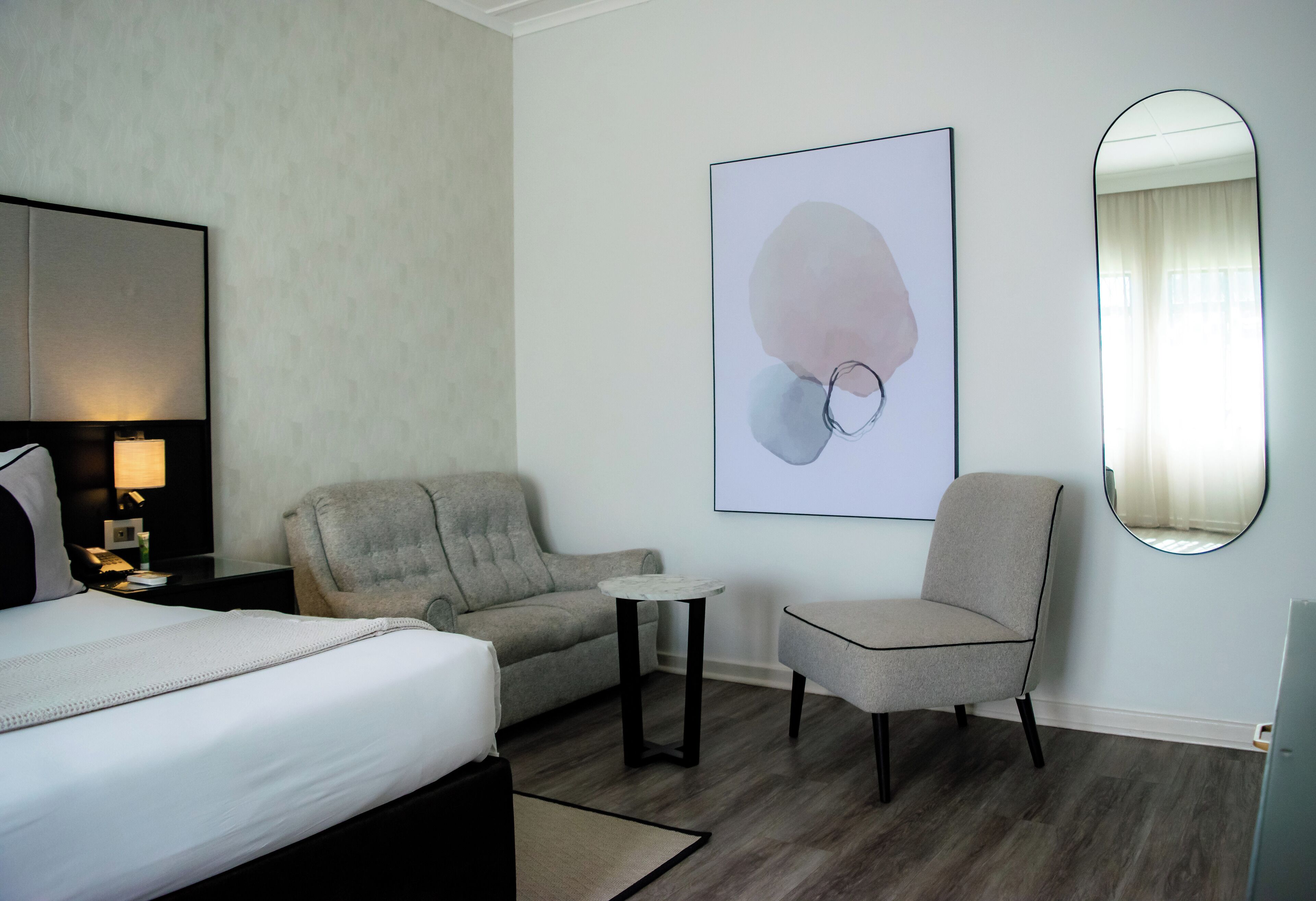 Una habitación de hotel con una cama, un sofá gris, una mesita, una silla, un espejo instalado en la pared y un cuadro abstracto enmarcado.