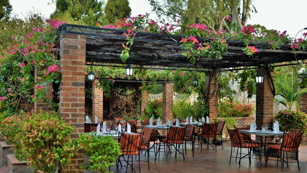 An outdoor patio with brick columns, tables, and chairs, surrounded by lush greenery and flowering plants. - Cresta Churchill (Bulawayo)