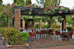 Un patio al aire libre con columnas de ladrillo, mesas y sillas, rodeado de abundante vegetación y plantas con flores.