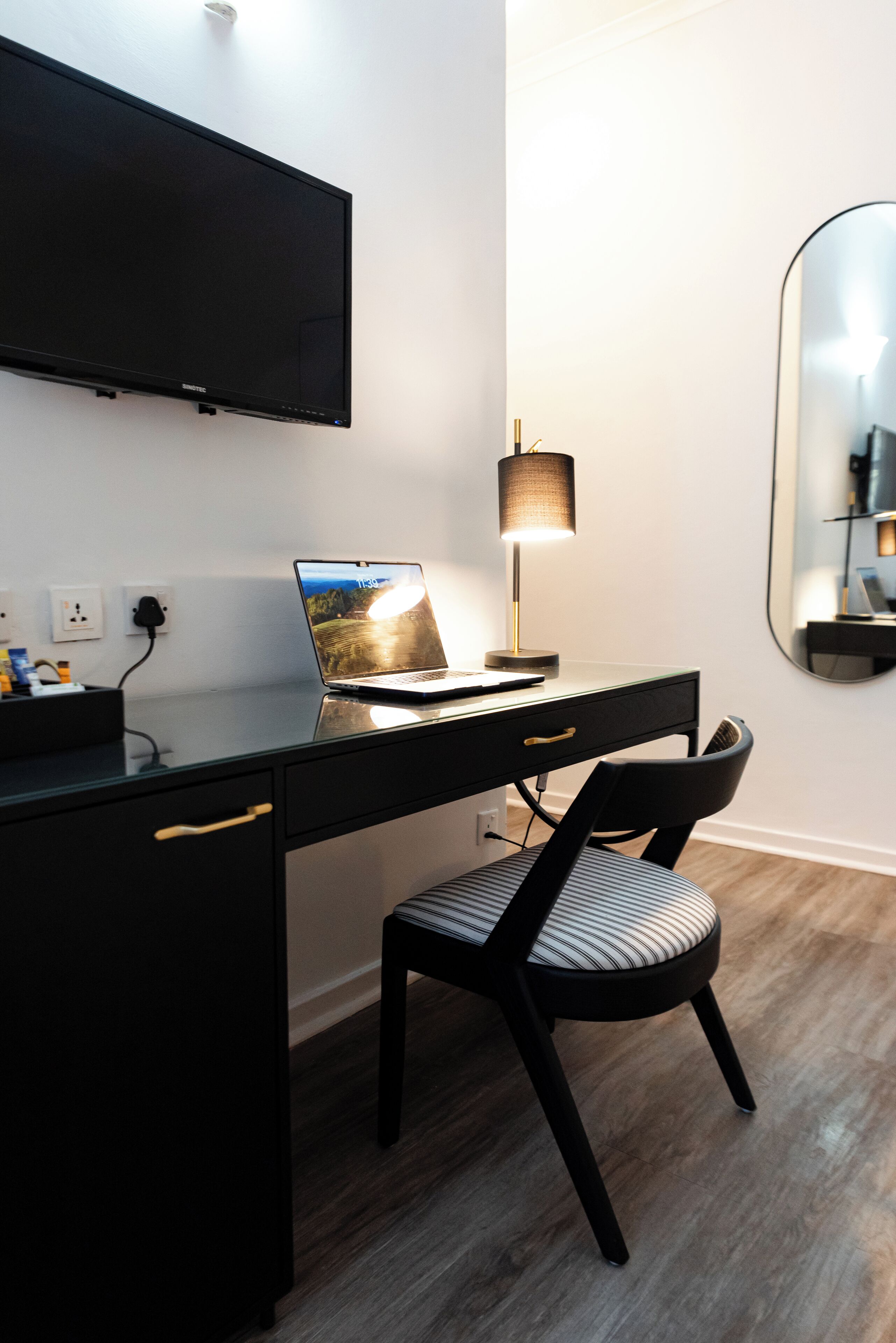 A modern room with a black desk, a laptop, a lamp, a chair, and a TV mounted on the wall.