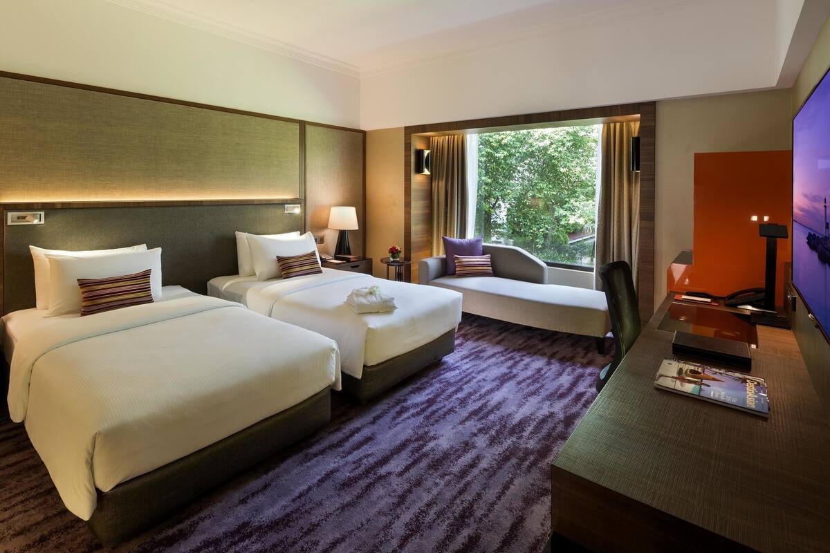 Habitación de hotel con dos camas, un escritorio, televisión y ventana con vista a zonas verdes.