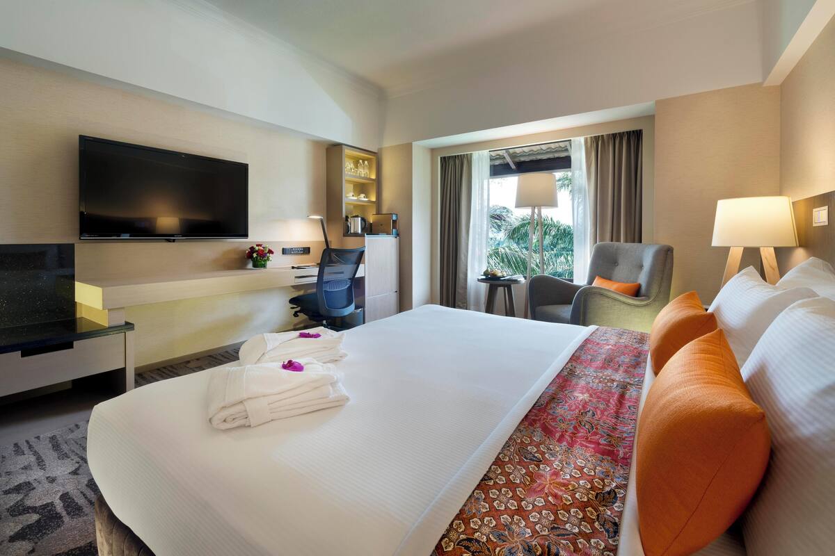 A hotel room with a bed, a TV, a desk with a chair, a small table, and a window with curtains.
