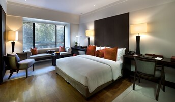 A modern hotel room with a large bed, a desk, a chair, a sofa, and a window with blinds.