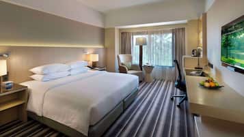 A hotel room with a bed, bedside tables, a desk with a lamp, a chair, a TV, and a fruit bowl.