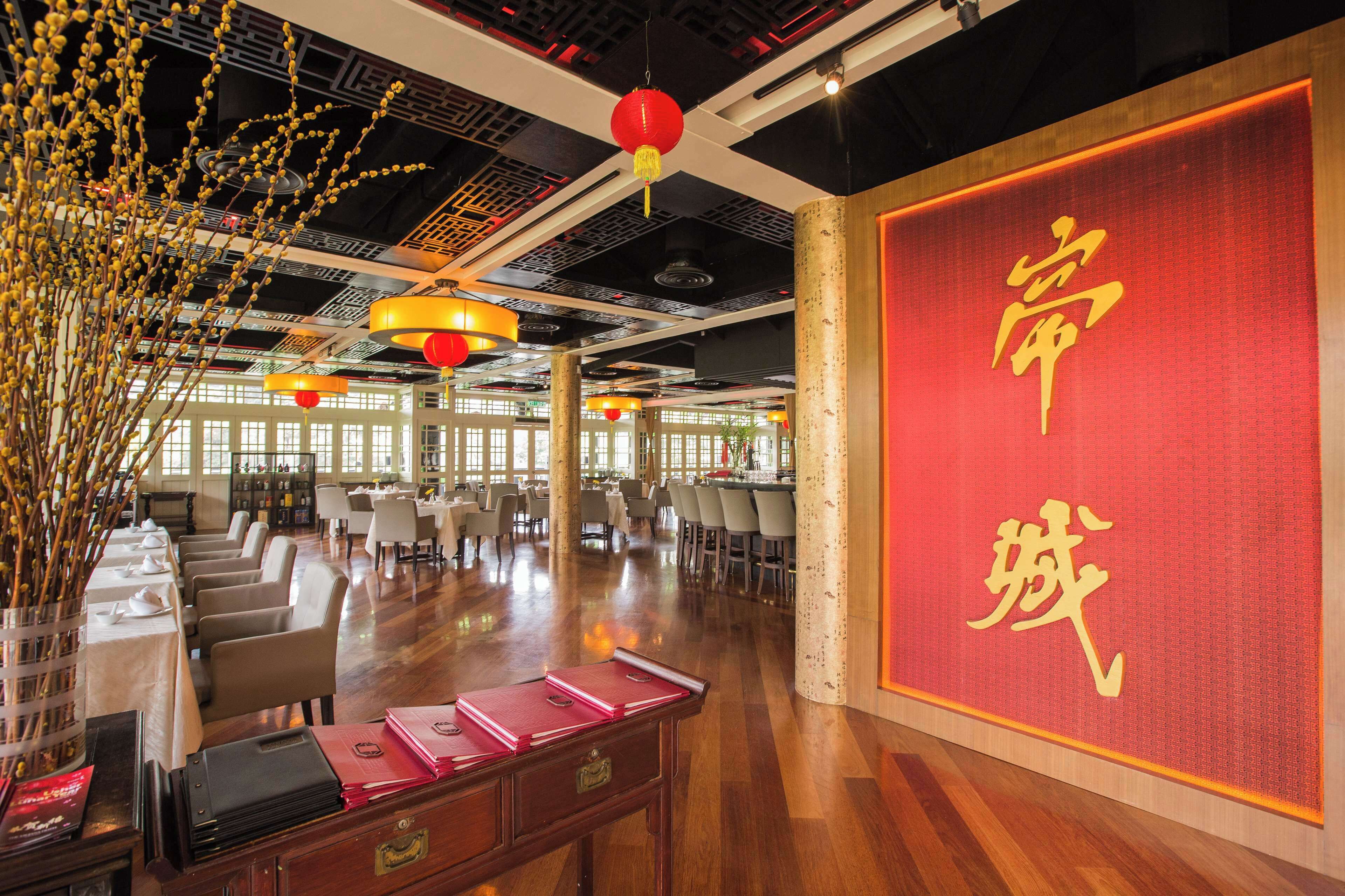 A spacious hotel lobby with a traditional Asian design, featuring a large red wall with golden characters, wooden furniture, and hanging red lanterns.