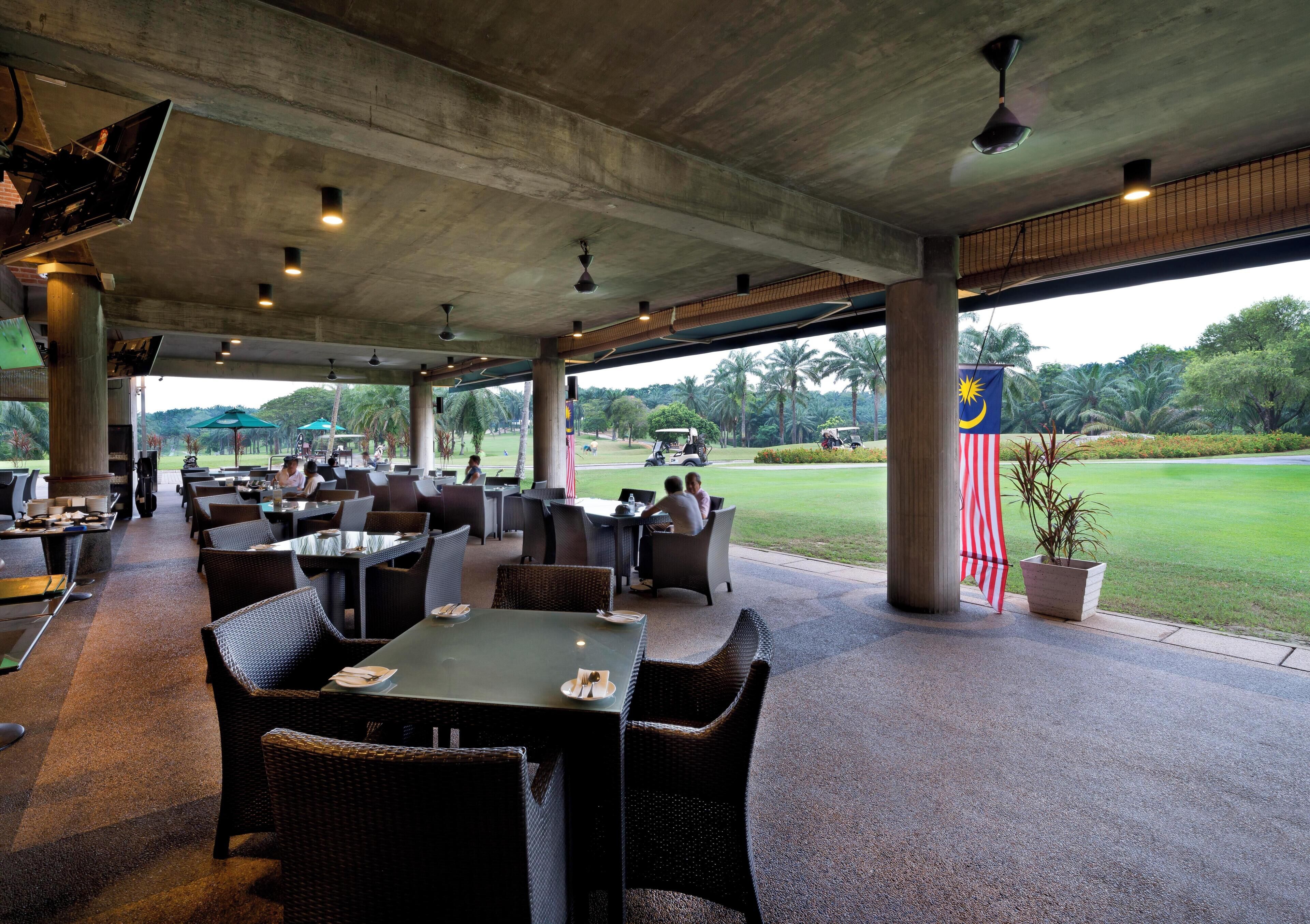 Un área de comedor al aire libre cubierta con mesas y sillas, vista a un campo de golf y una bandera de Malasia.