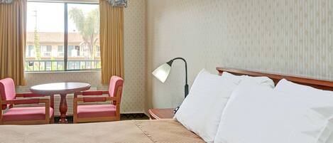 Habitación de hotel con cama, una mesa redonda, dos sillas, una lámpara y una ventana con cortinas.