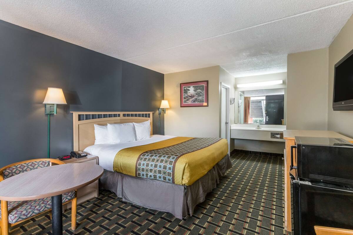 A hotel room with a bed, bedside lamps, a small table, a chair, a TV, and a window.