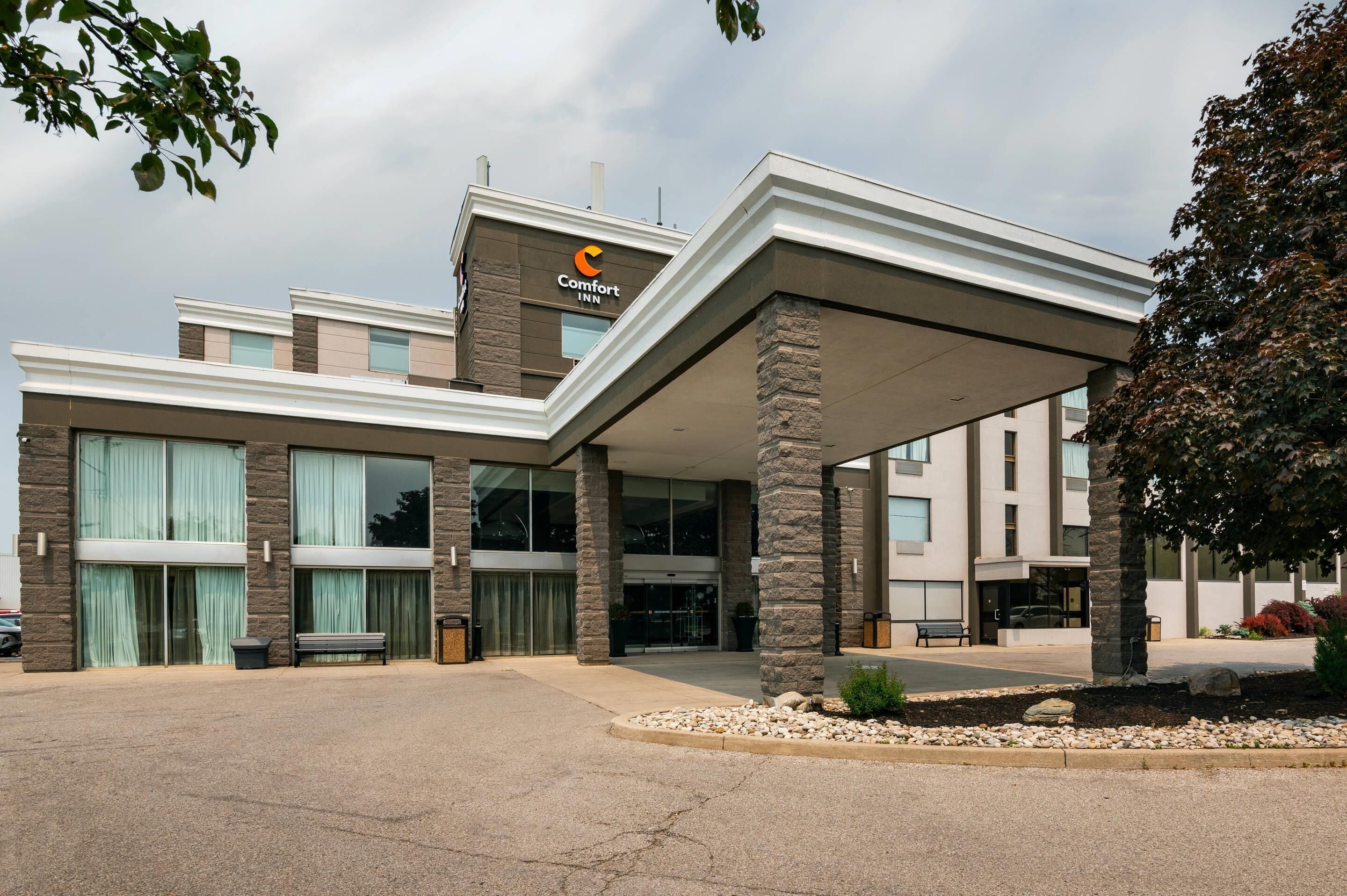 A Comfort Inn hotel with a modern design, featuring a covered entrance, stone pillars, and a landscaped area with a bench.