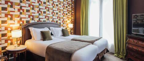 A hotel room with a patterned wall, a large bed with multiple pillows, a bedside table with a lamp, and a television on a dresser.