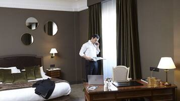 A hotel room with a bed, a desk, a chair, a lamp, and a window with curtains.