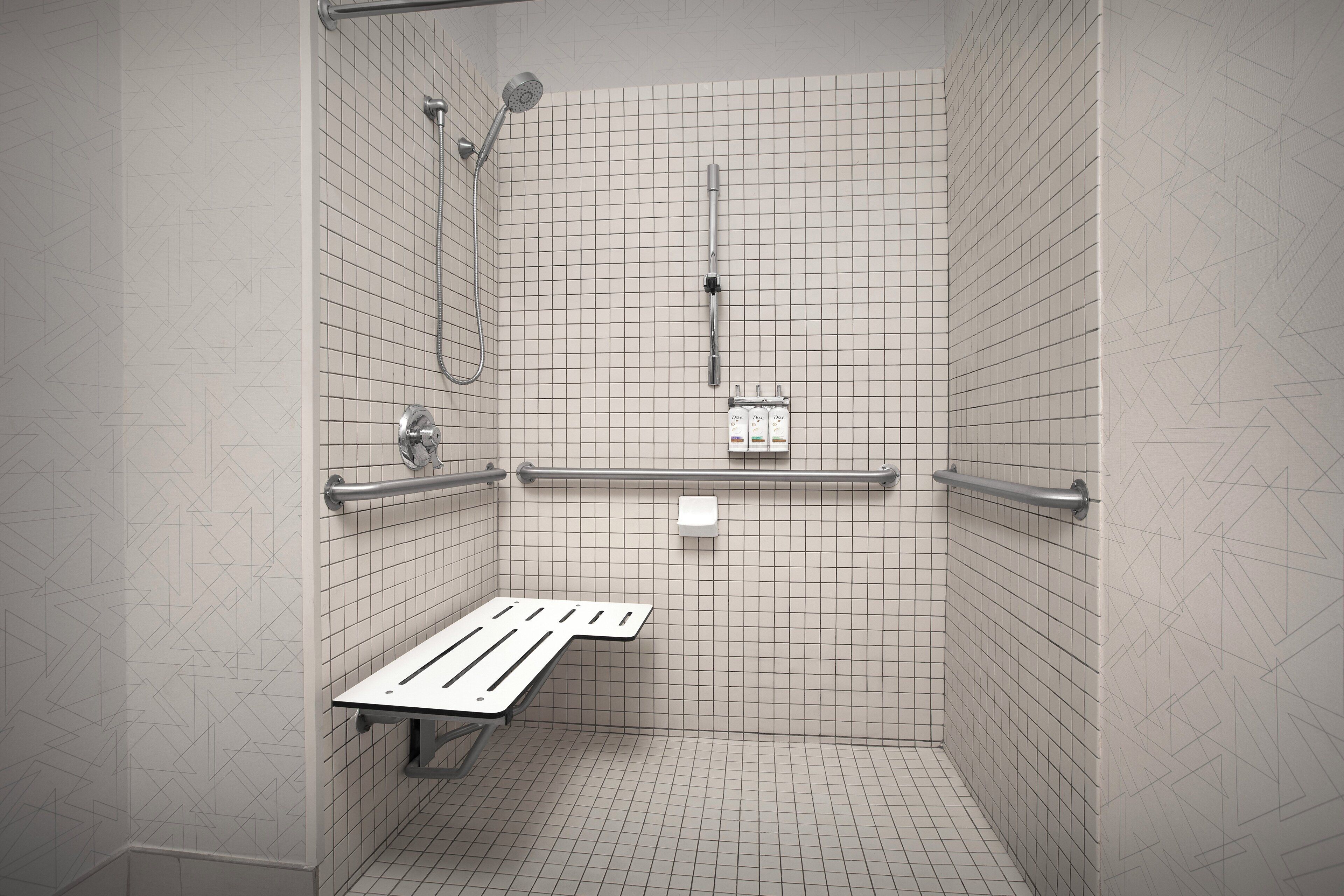 A small, tiled shower stall with a bench, grab bars, and a handheld showerhead.