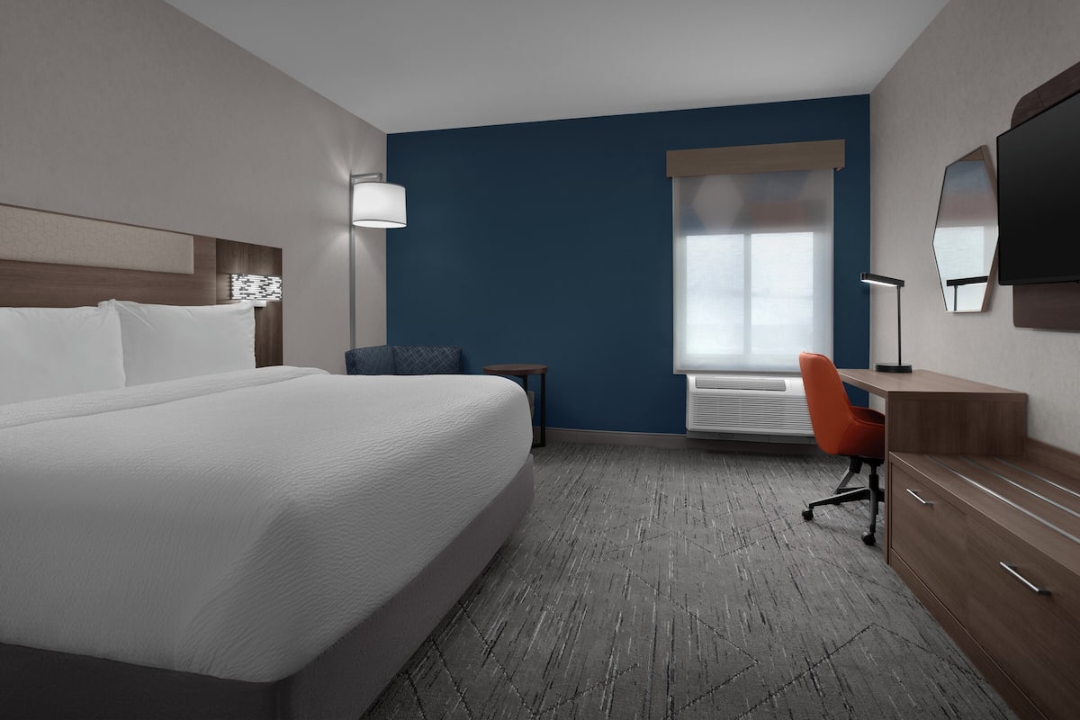 A hotel room with a large bed, a desk with a chair, a flat-screen TV, and a window with blinds.