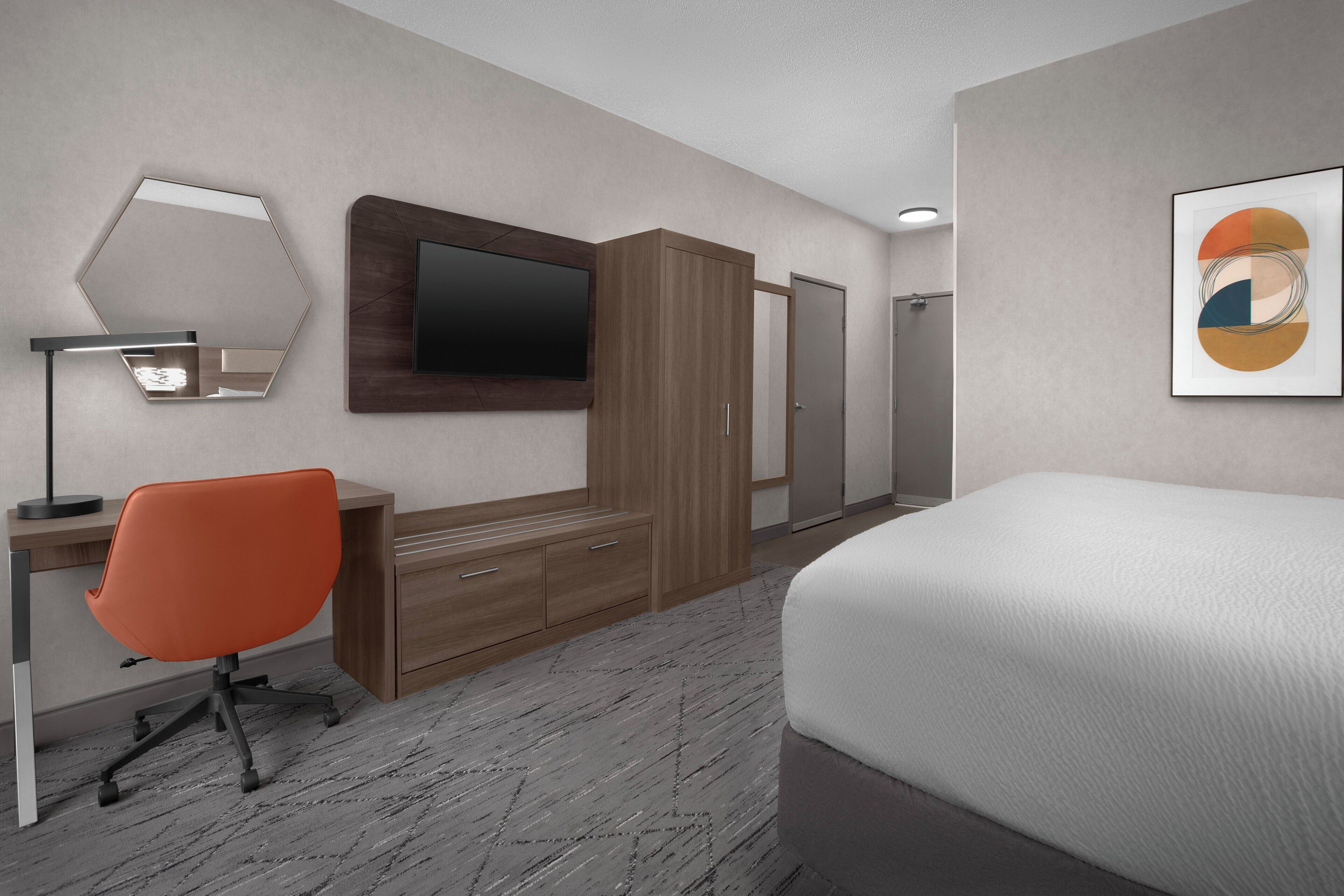 A modern hotel room with a bed, a desk with a computer, an orange office chair, a TV on the wall, and a wardrobe.