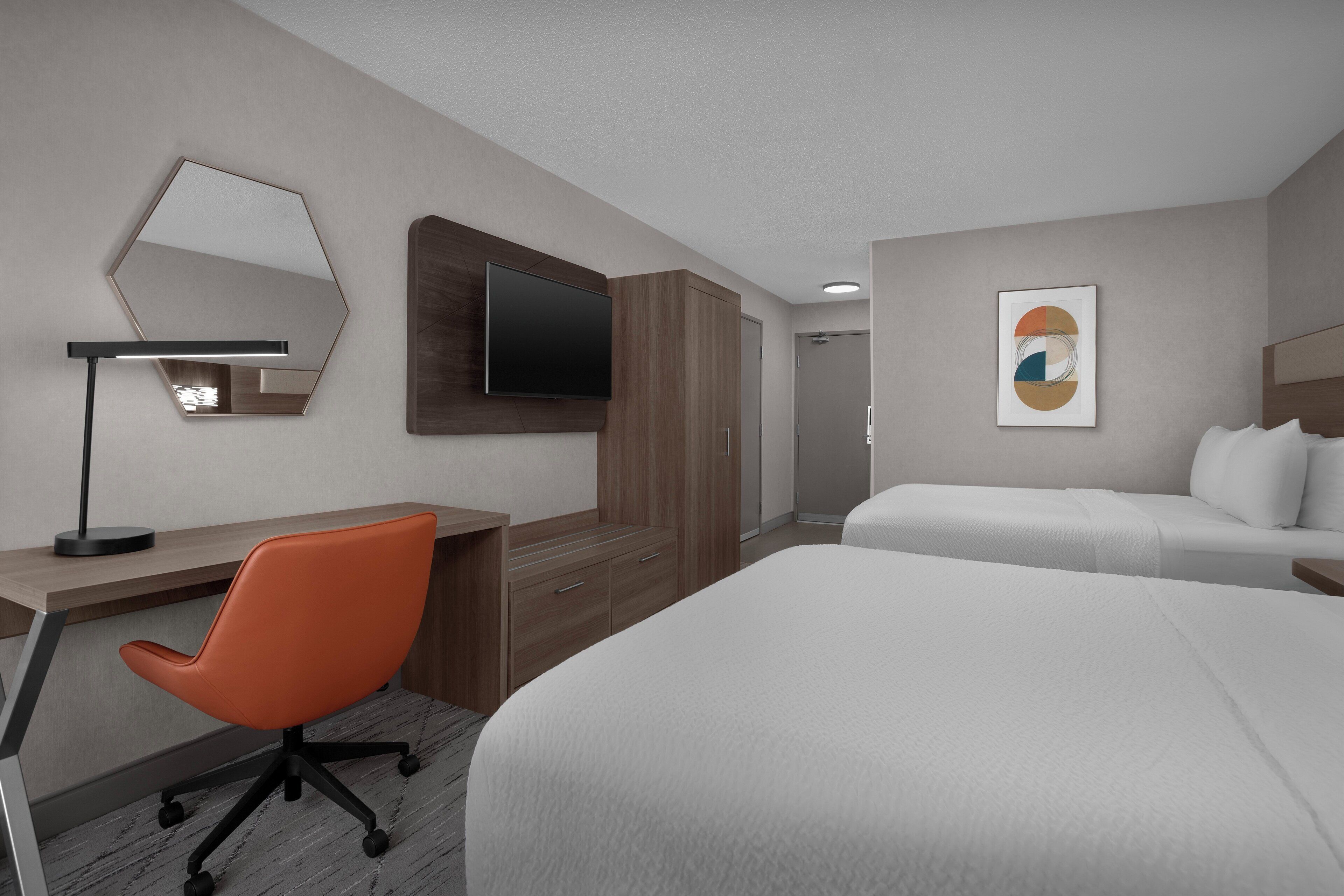 A hotel room with a desk, an orange office chair, a bed, a TV mounted on the wall, and a framed artwork.