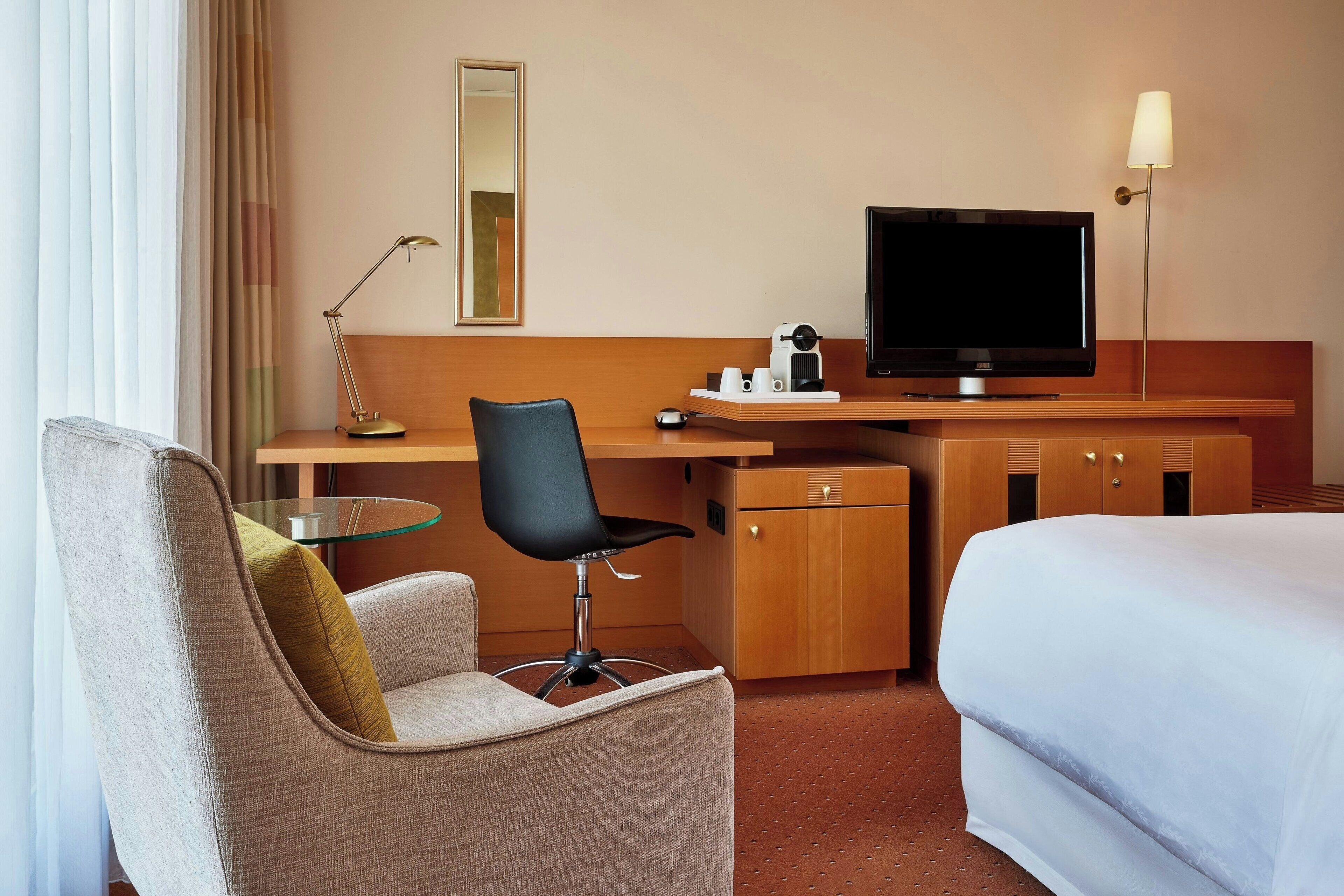 Ein Hotelzimmer mit einem Bett, einem Schreibtisch mit Fernseher, einem Sessel, einer Lampe und einem Spiegel.