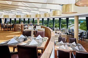 A modern restaurant with high ceilings, large windows, and a view of the cityscape. - Sheraton Duesseldorf Airport Hotel (Düsseldorf)