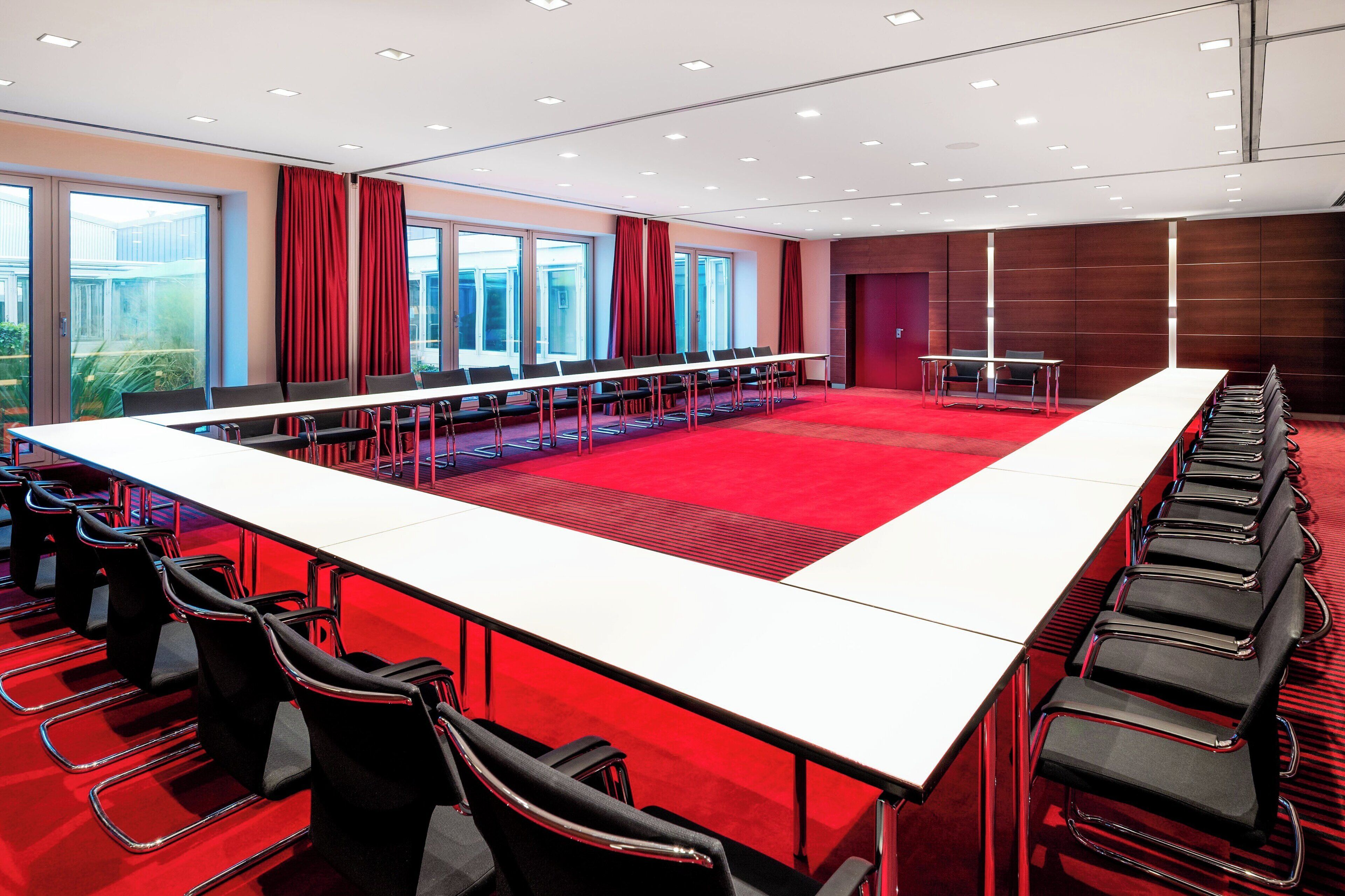 Un salón de conferencias con una larga mesa, filas de sillas y ventanas grandes con cortinas rojas.