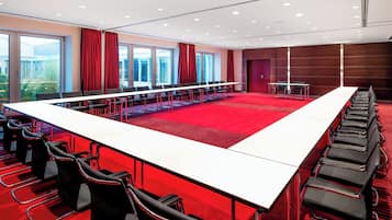 Un salón de conferencias con una larga mesa, filas de sillas y ventanas grandes con cortinas rojas.