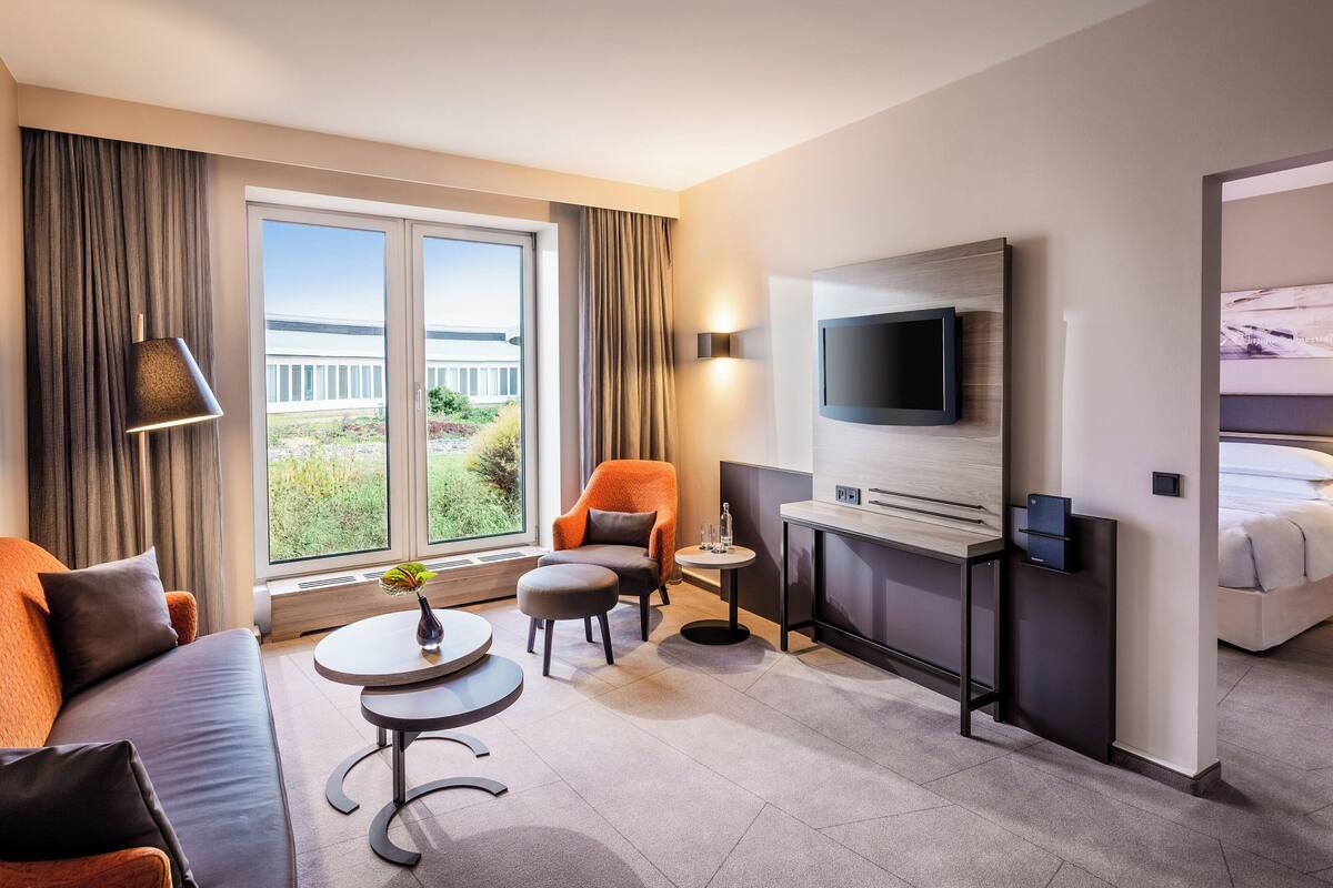 Habitación de hotel con sofá, sillón, mesita redonda y televisor montado en la pared.