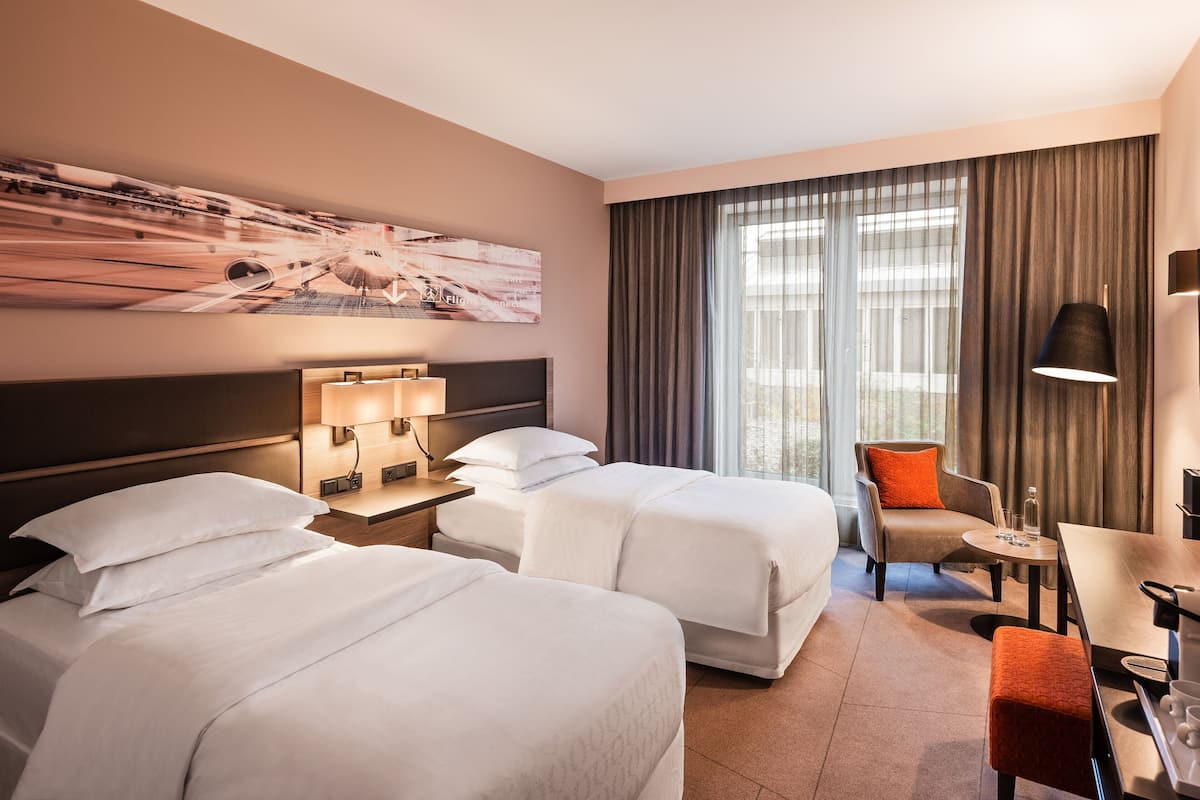 Habitación de hotel con dos camas, una silla, una mesa y un amplio ventanal con cortinas.