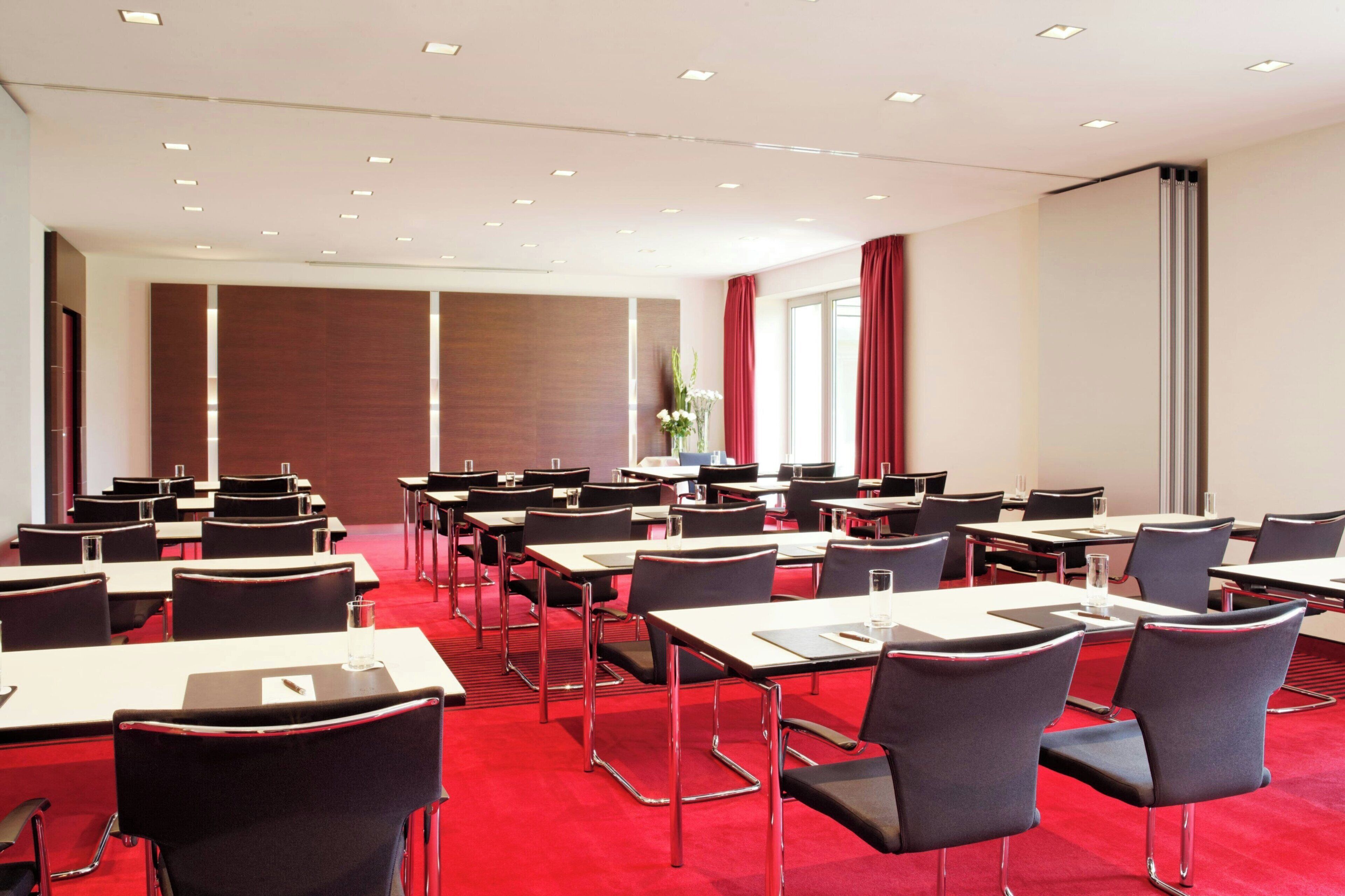A conference room with rows of tables and chairs, a red carpet, and a large window with curtains.