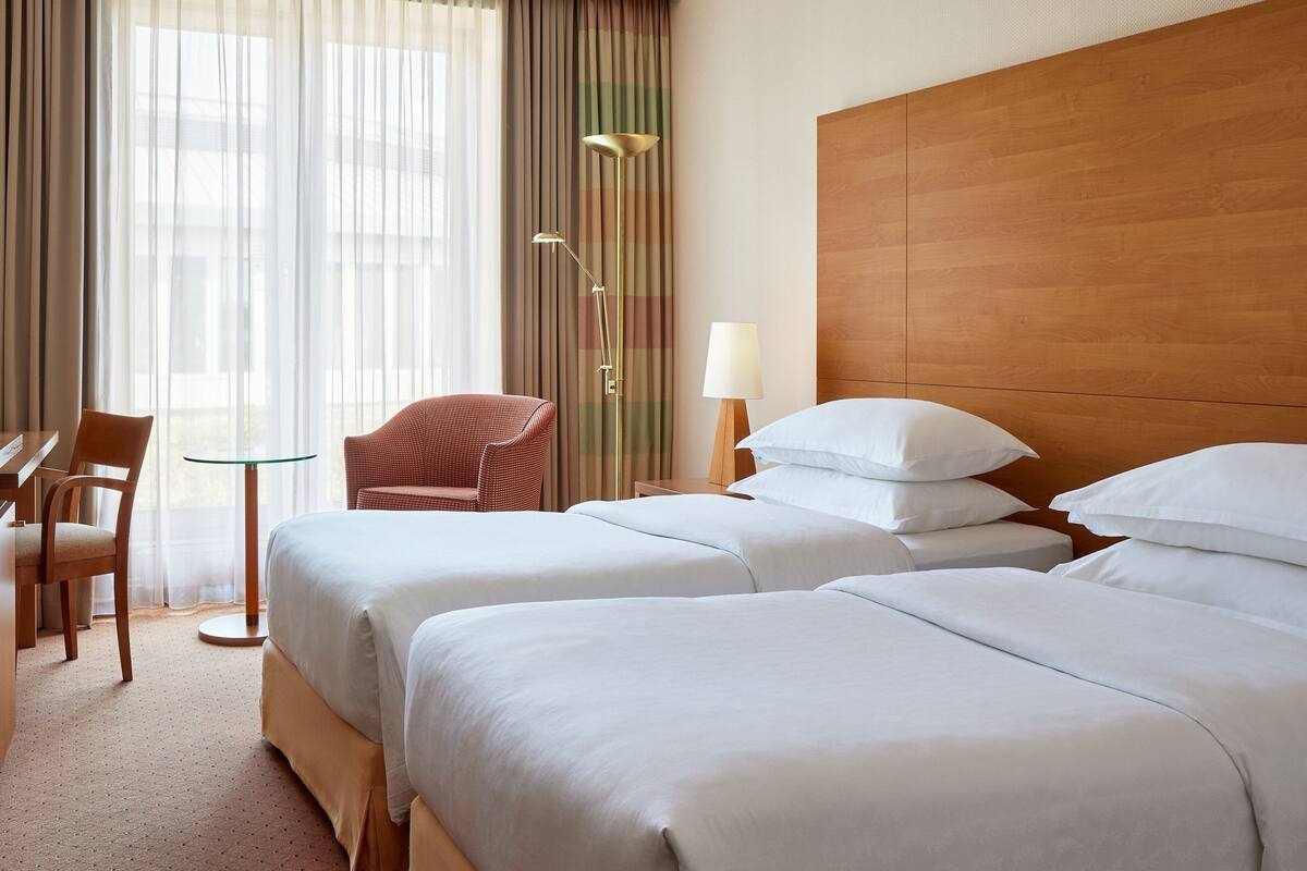 Habitación de hotel con dos camas, un escritorio, una silla, una lámpara y vistas al exterior a través de cortinas translúcidas.