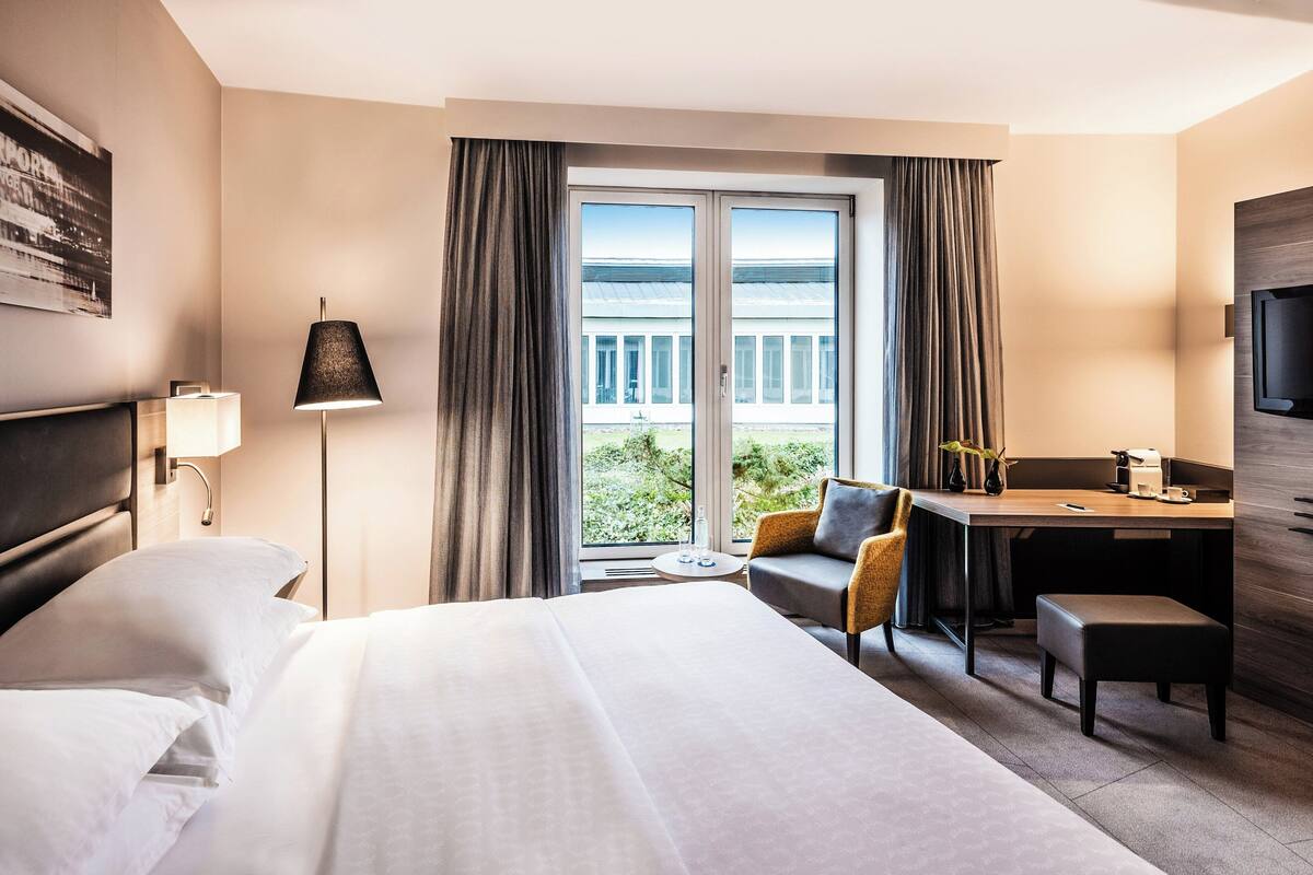 Habitación de hotel con una cama grande, un escritorio con silla, un televisor y un balcón con cortinas.