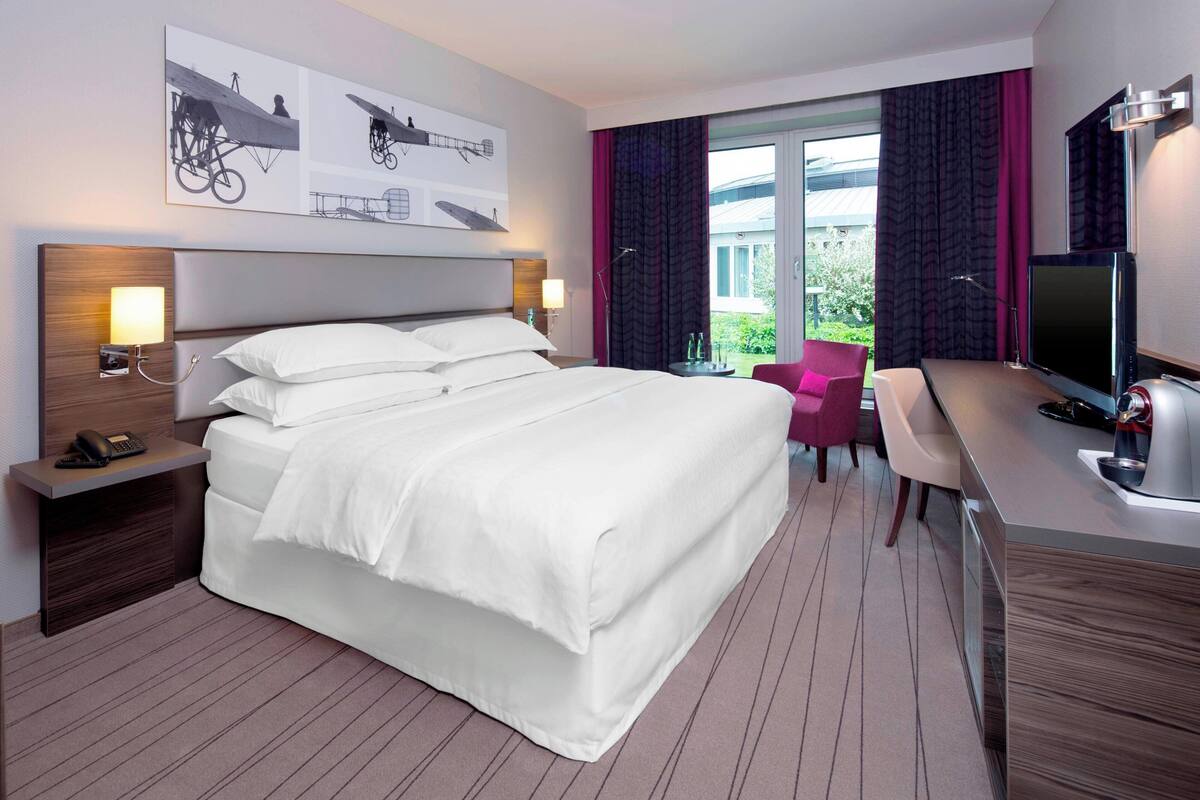 Habitación de hotel con una cama grande, un escritorio con silla, televisión y ventana con cortinas.