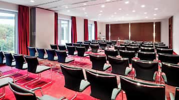 Un salón de conferencias con filas de sillas, un atril y ventanas grandes con cortinas.
