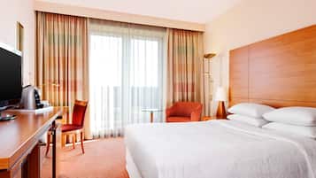 Quarto de hotel com cama, escrivaninha, televisão, cadeira, abajur e cortinas.